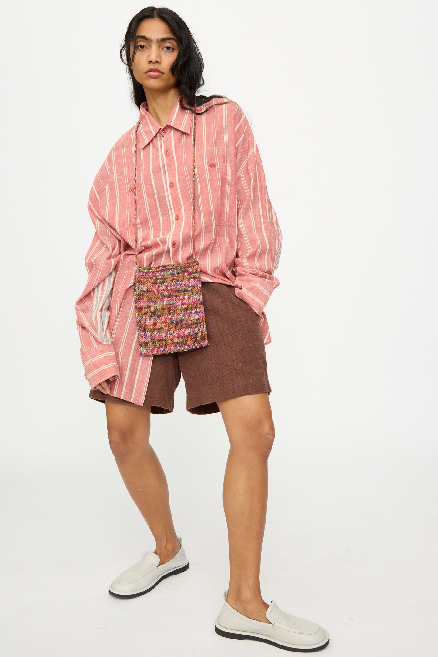 A person in a pink striped oversized shirt, brown shorts, white slip-on shoes, and the 11.11 Silk Knitted Bag in Multi stands against a plain white background.