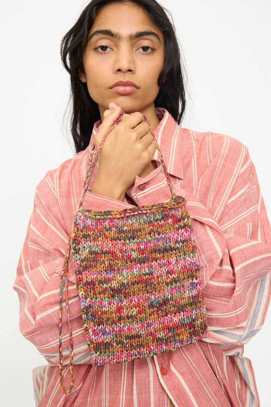 A person in a pink striped shirt holds the 11.11 Silk Knitted Bag in Multi, a limited edition crossbody with a braided strap, against a plain background.