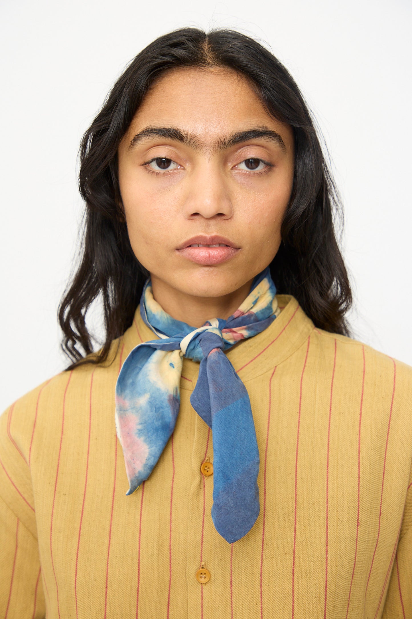 A person with long dark hair wears a yellow shirt with red stripes and an 11.11 Silk Scarf in Indigo tied at the neck, standing against a plain light background.