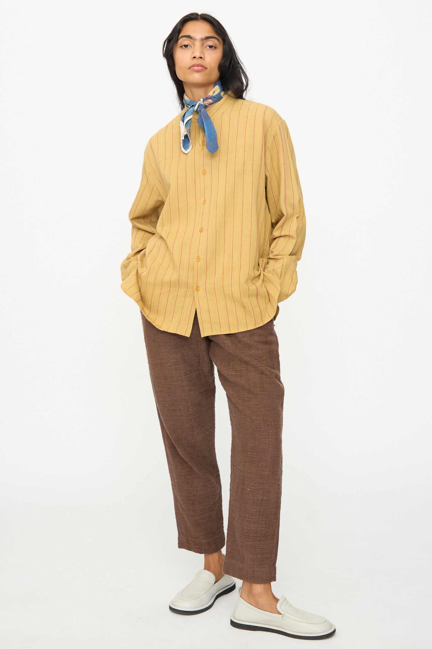 A person stands against a white background wearing a yellow pinstripe button-up shirt, brown pants, white slip-on shoes, and the 11.11 Silk Scarf in Indigo.