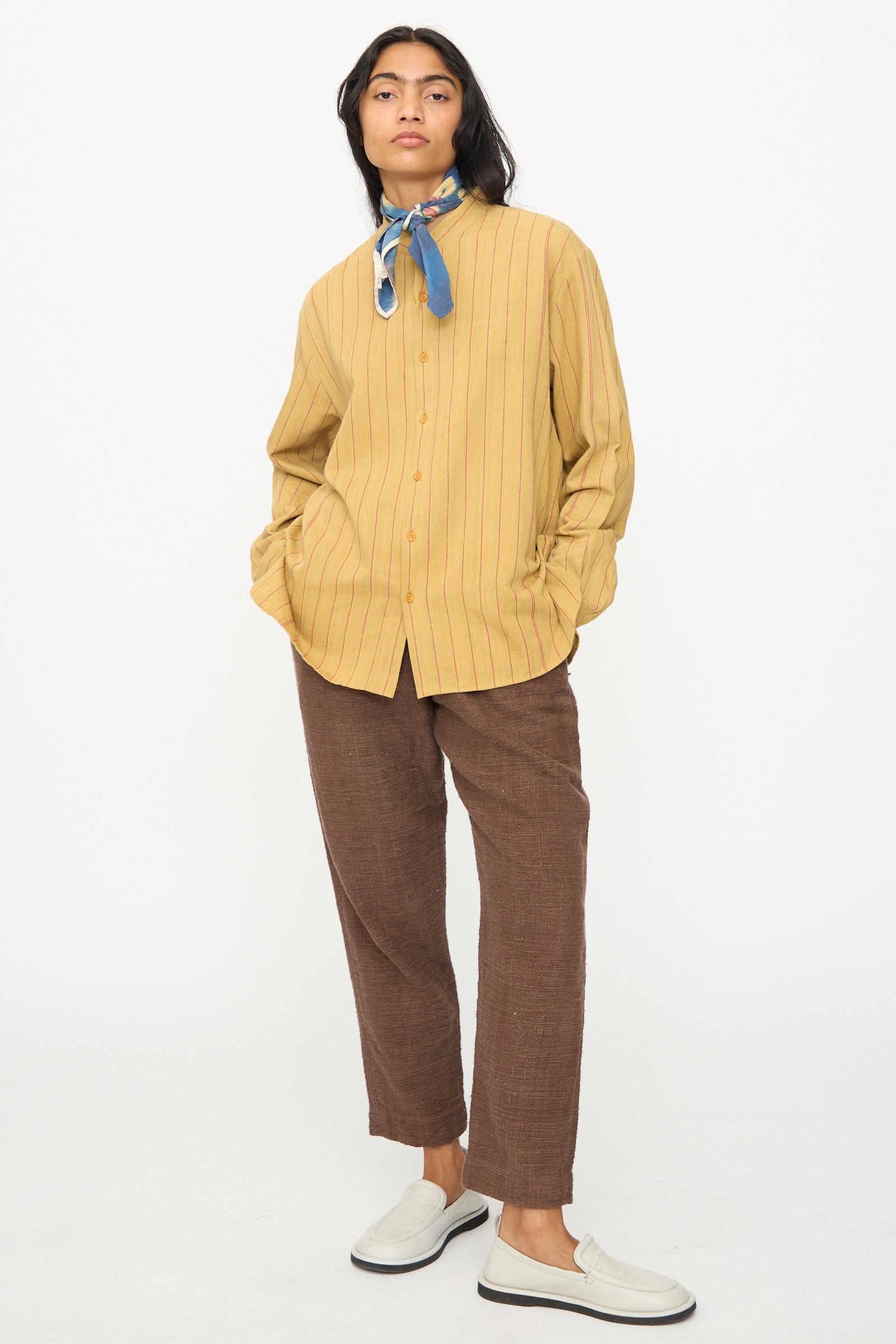 A person stands against a white background wearing a yellow pinstripe button-up shirt, brown pants, white slip-on shoes, and the 11.11 Silk Scarf in Indigo.