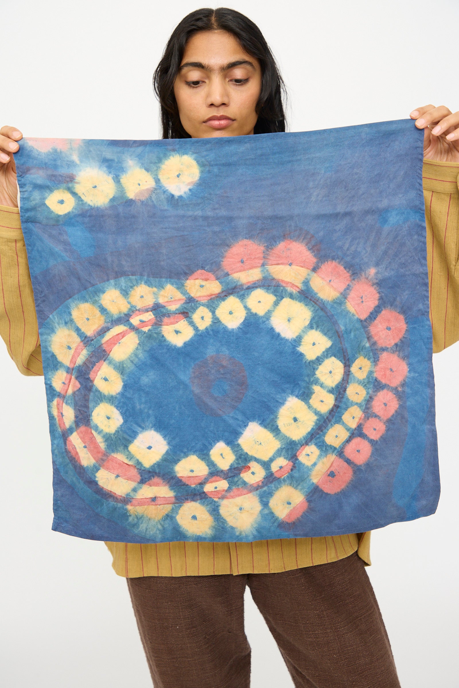 A person holds up the 11.11 Silk Scarf in Indigo, featuring circular yellow and orange tie-dye patterns.