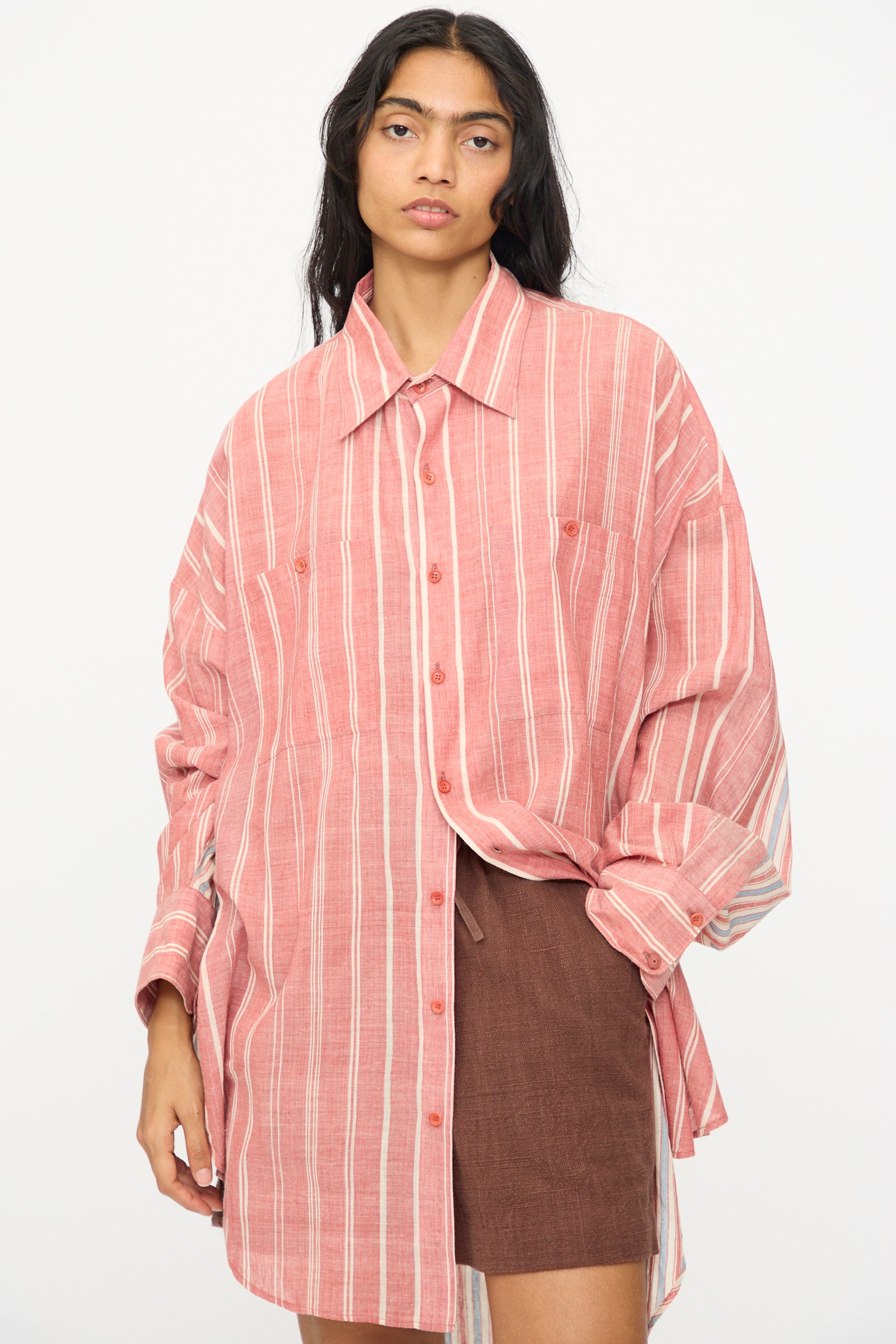 A person wears the 11.11 Cotton Stripe Shirt in Multi—an oversized, long-sleeve pink and white striped button-up—over brown shorts, standing against a plain light background.