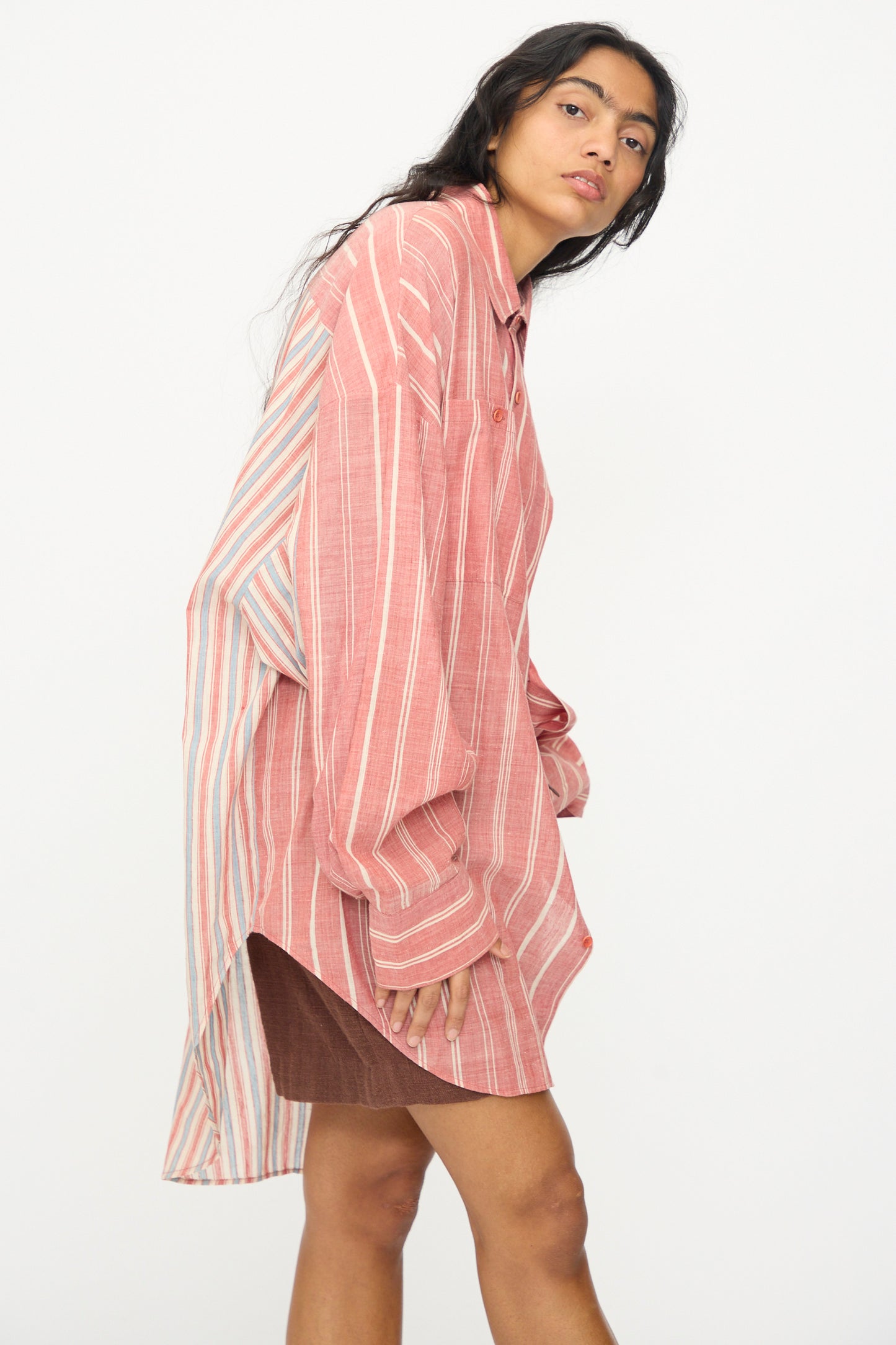 A woman wears the 11.11 Cotton Stripe Shirt in Multi—an oversized pink and white striped button-up with a high-low hem—paired with a short brown skirt, standing against a plain white background. Made in India.
