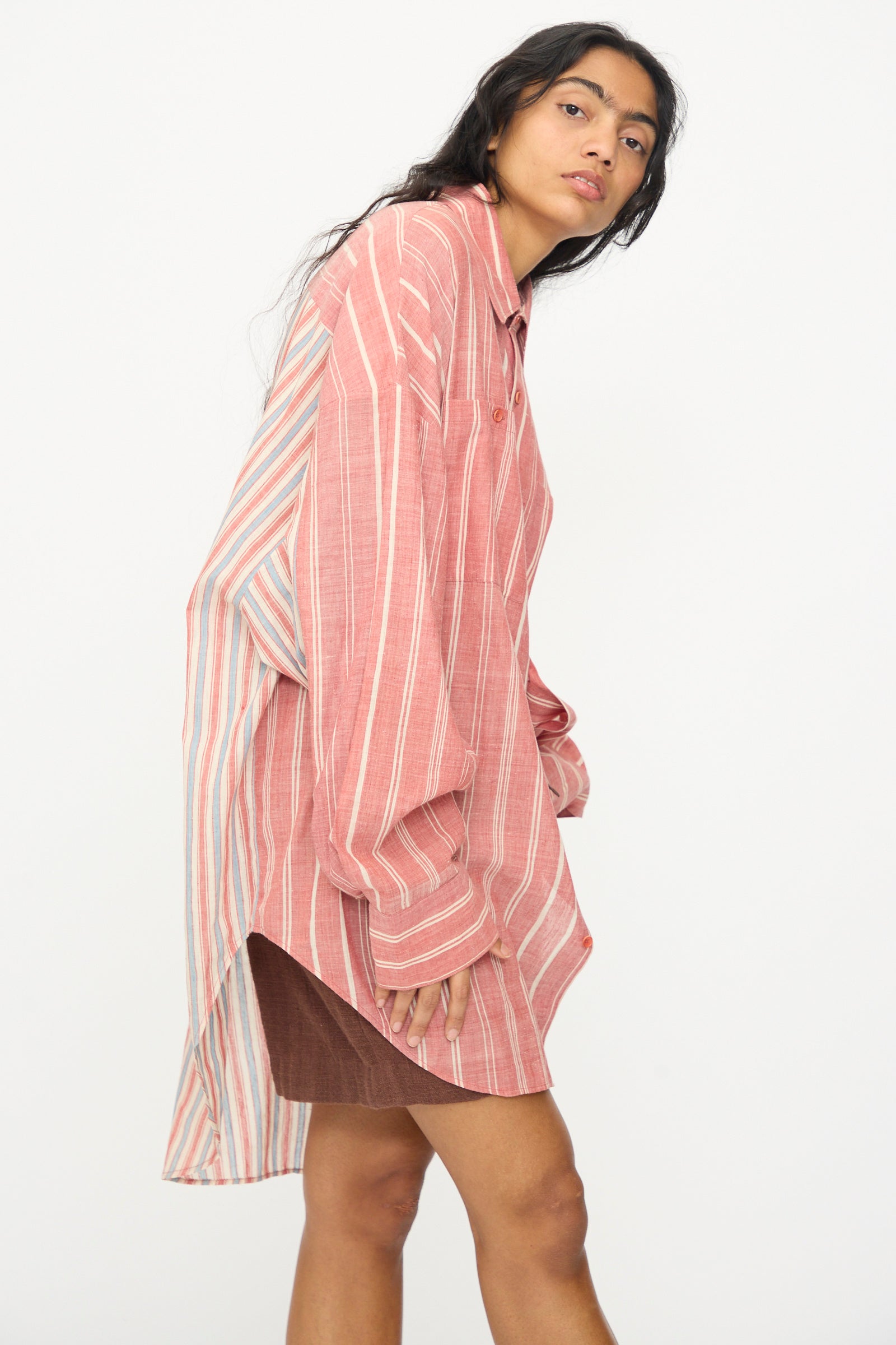 A woman wears the 11.11 Cotton Stripe Shirt in Multi—an oversized pink and white striped button-up with a high-low hem—paired with a short brown skirt, standing against a plain white background. Made in India.