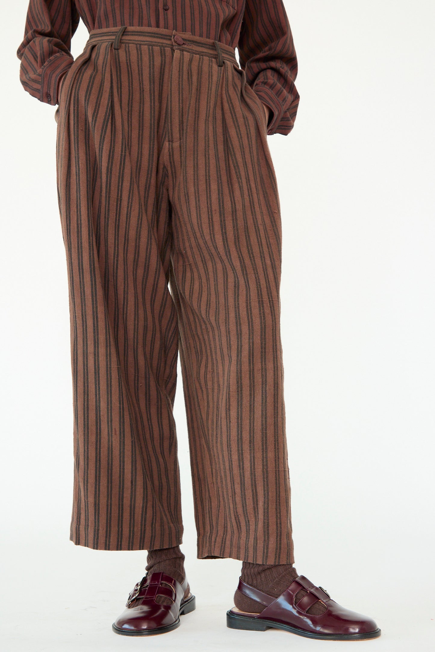 A person sports 11.11's Tel Trouser in Burnt Umber Stripe and a matching shirt, both naturally dyed. The look is completed with brown socks and burgundy shoes, exuding effortless style.