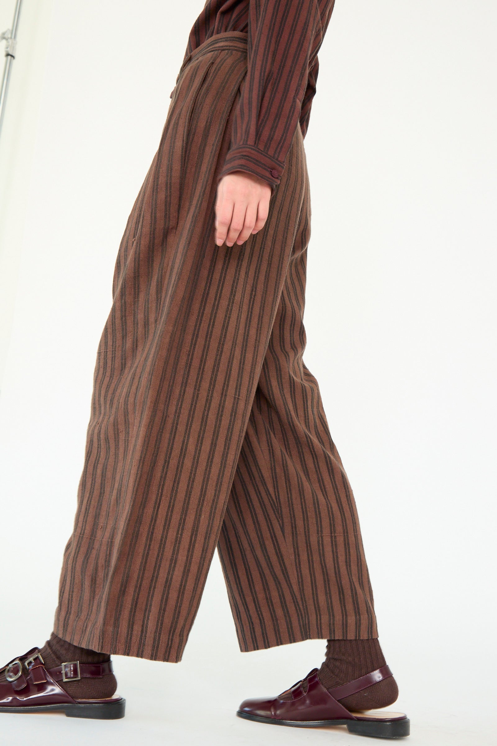 A person models the Tel Trouser in Burnt Umber Stripe by 11.11 against a light background. They complete the look with a maroon striped long-sleeve shirt, matching maroon shoes, and coordinating brown socks for a cohesive ensemble.