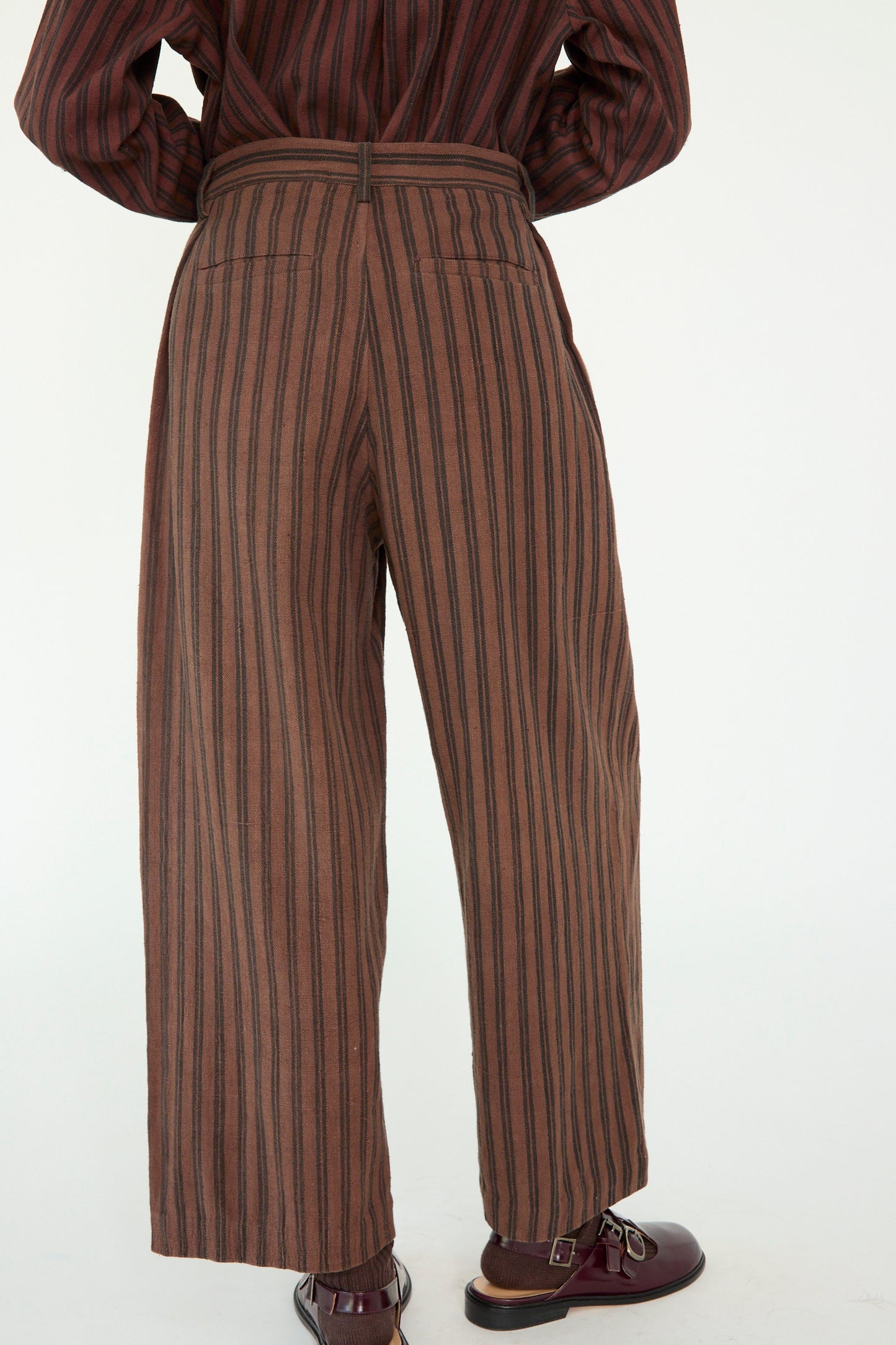 A person is seen from behind wearing 11.11's Tel Trouser in Burnt Umber Stripe, paired with dark brown shoes.