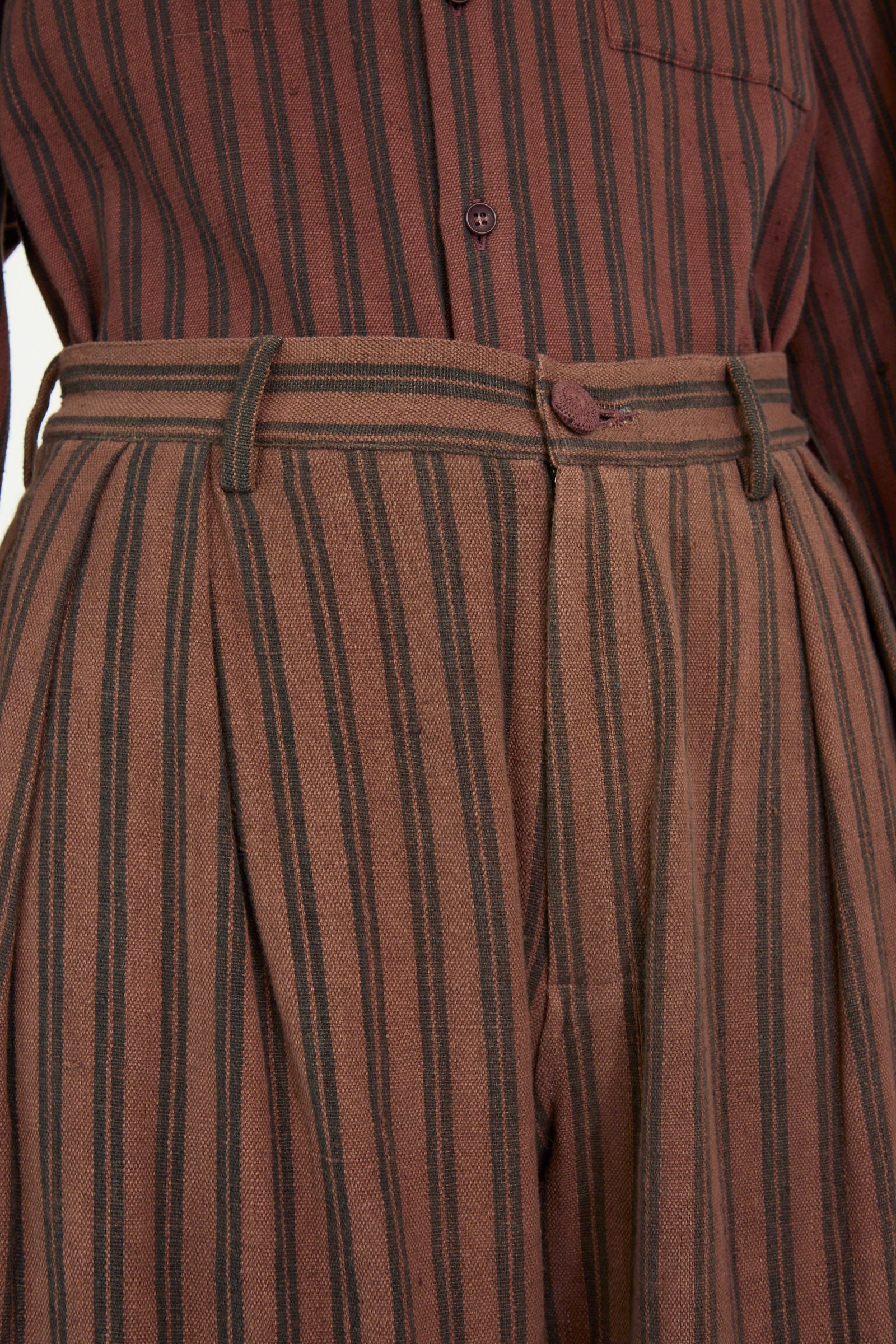 Close-up of 11.11's Tel Trouser in Burnt Umber Stripe, featuring a buttoned long-sleeve top and striped high-waisted pants with belt loops, crafted using a natural dye process.