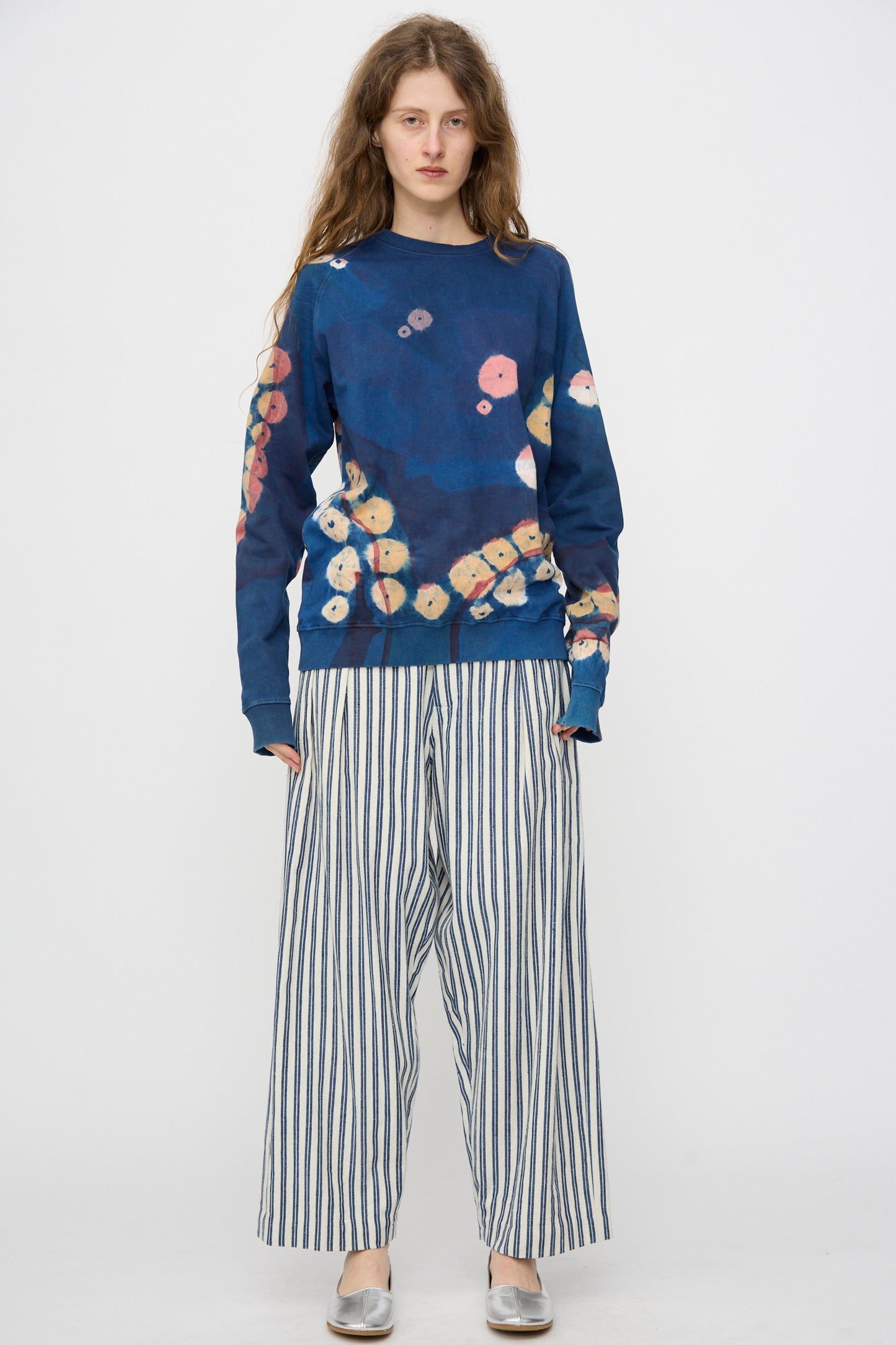 A woman stands against a plain background wearing a blue sweatshirt with a colorful pattern and 11.11 Tel Trouser in Medium Indigo Stripe, paired with silver flat shoes.