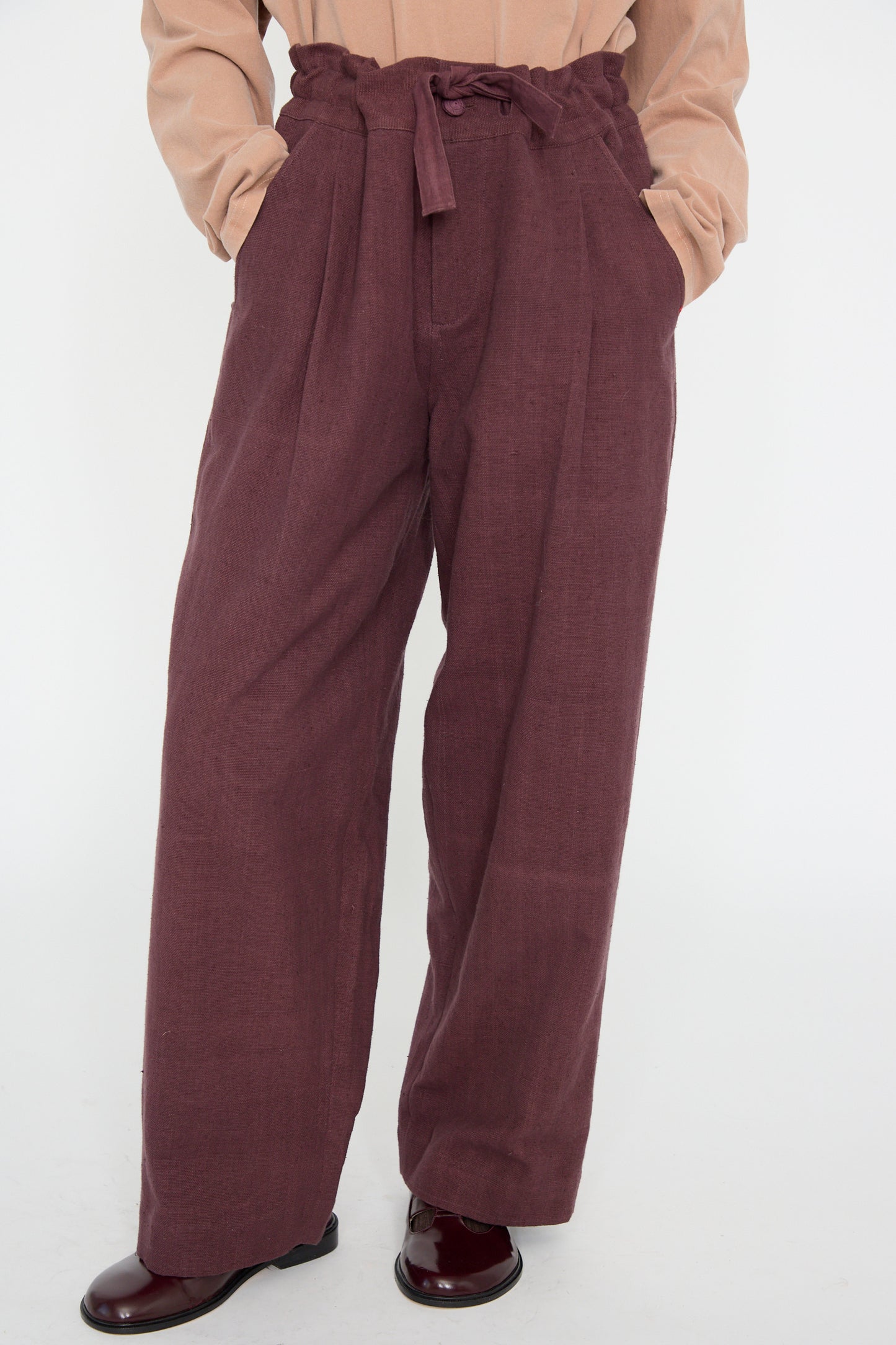 Person wearing relaxed-fit Zed Trouser in Purple Red from 11.11, with a woven cotton tie belt and hands in pockets, paired with a beige top and brown shoes.