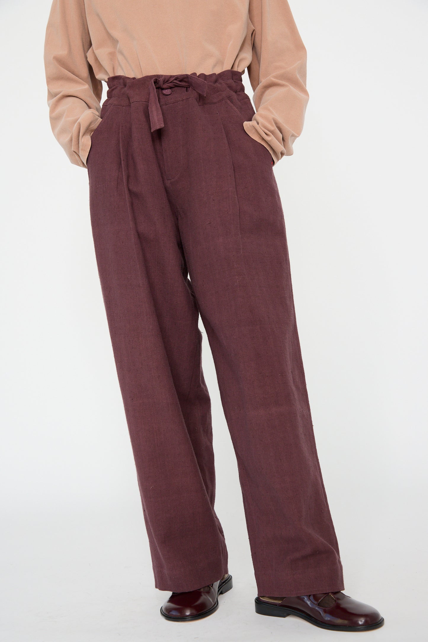 A person wears 11.11's Zed Trouser in Purple Red, featuring a high-waisted, relaxed fit with a drawstring waist, hands in pockets. The trousers are styled with a beige top against a plain backdrop.