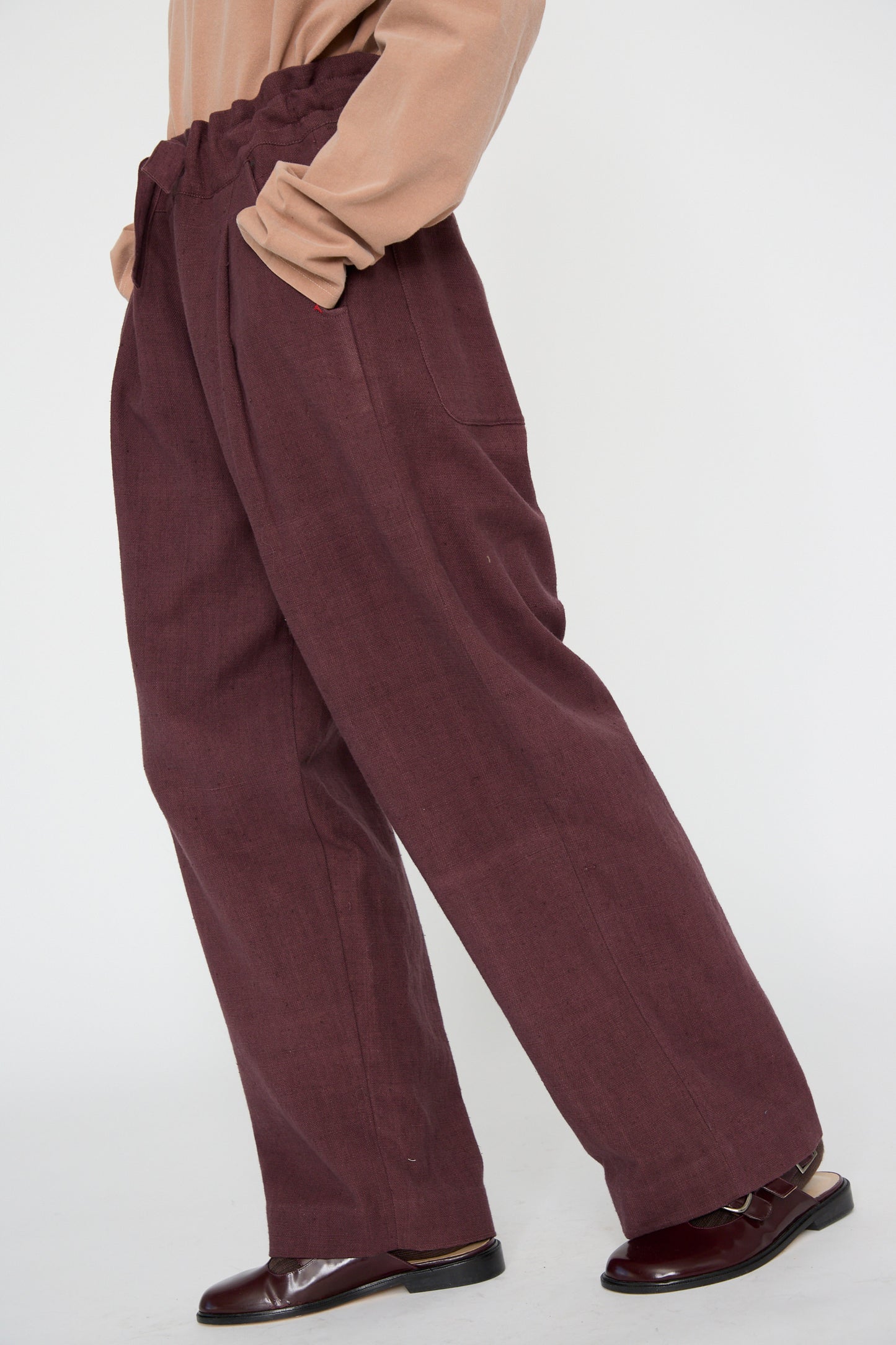 A person is wearing the 11.11 Zed Trouser in Purple Red with a drawstring waist and hands in pockets, paired with a beige top and brown shoes, set against a plain background highlighting the relaxed fit of this woven cotton ensemble.