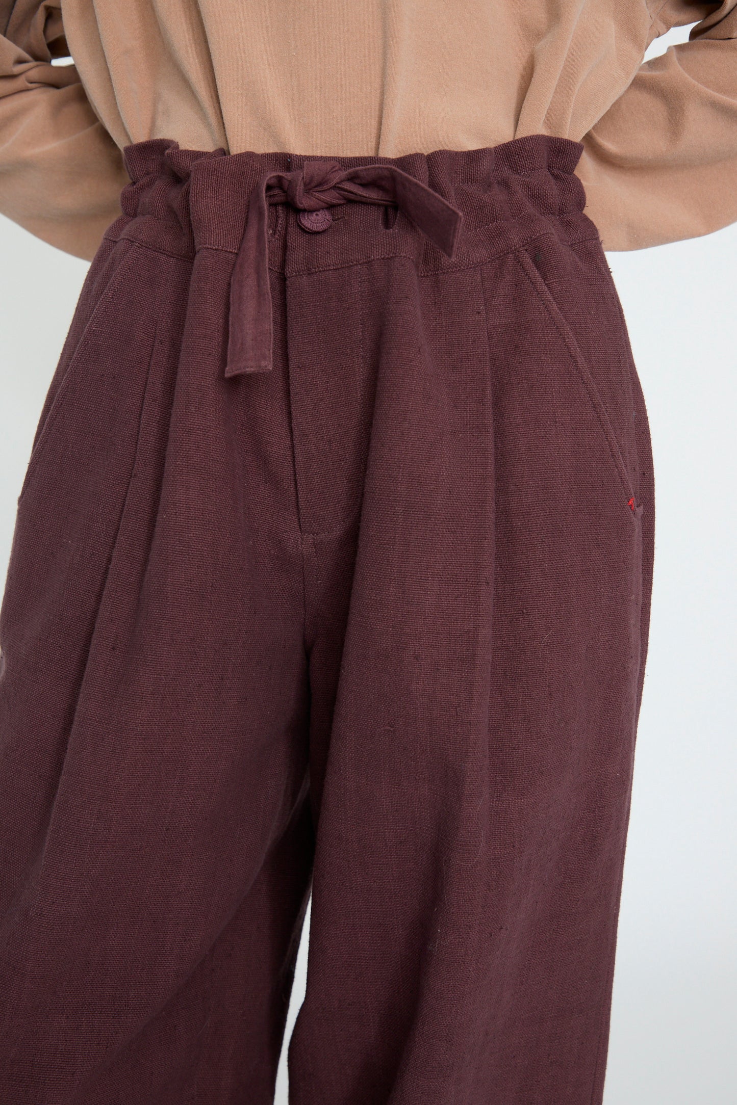 A person wearing 11.11's Zed Trouser in Purple Red, high-waisted woven cotton pants with a fabric tie belt, paired with a beige top.