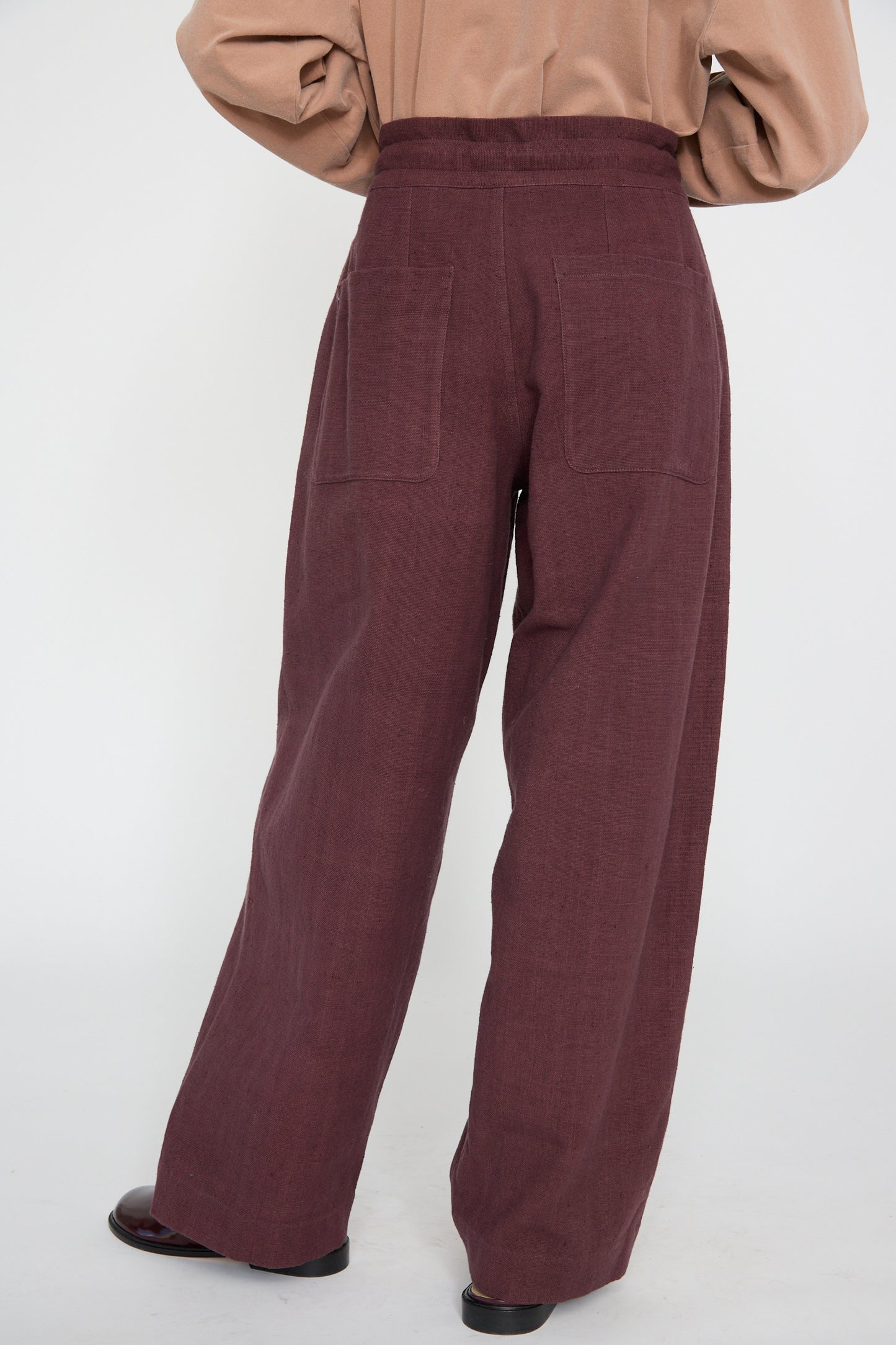 A person stands facing away, wearing the Zed Trouser in Purple Red by 11.11, featuring a relaxed fit with a high waist and wide legs. The stylish design includes woven cotton fabric and a comfortable drawstring waist.