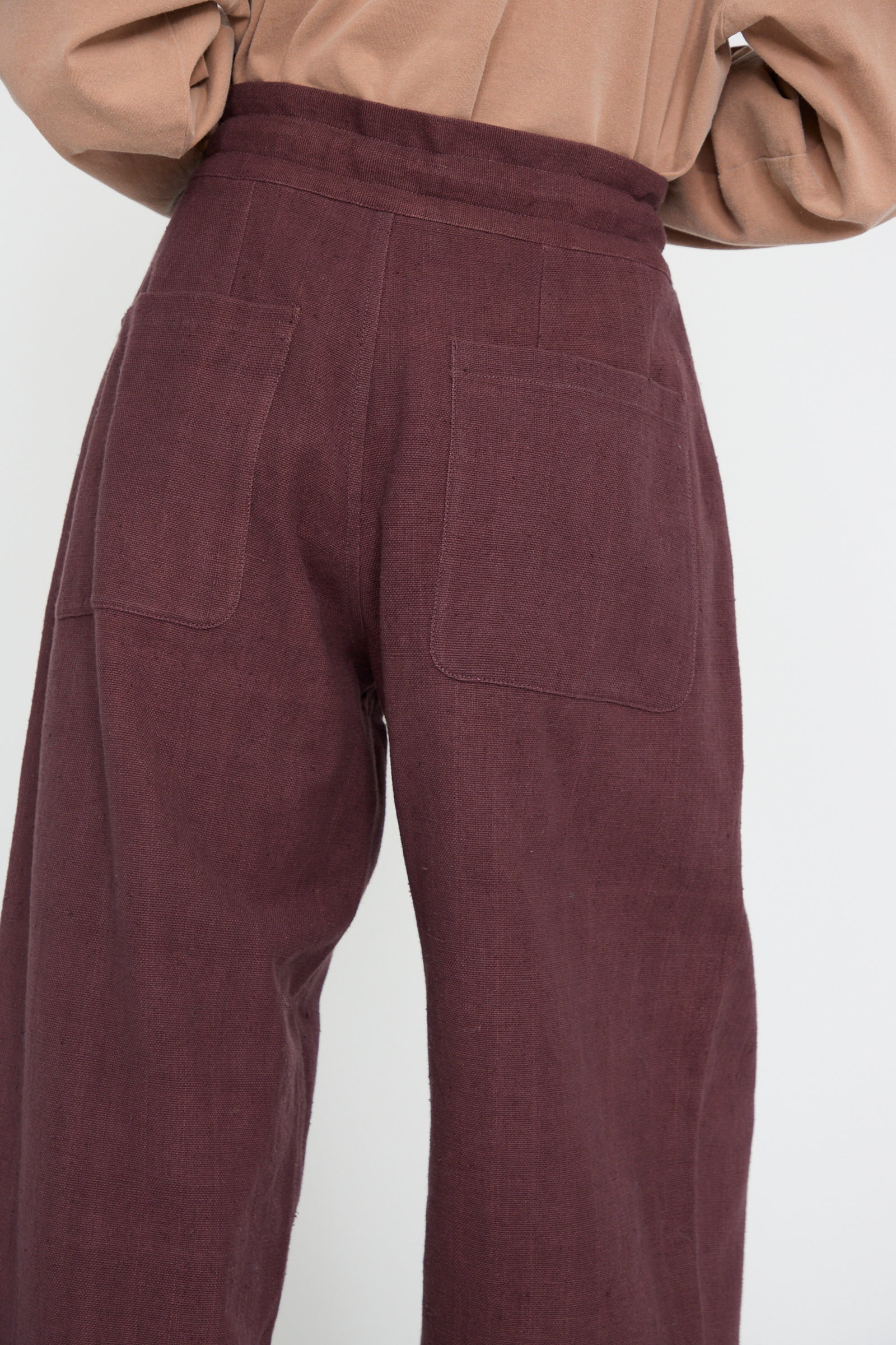 Against a light background, a person is seen in 11.11's Zed Trouser in Purple Red, boasting a relaxed fit with back pockets, high-waisted design, and crafted from woven cotton for added texture. They pair it with a beige top for an understated elegant look.