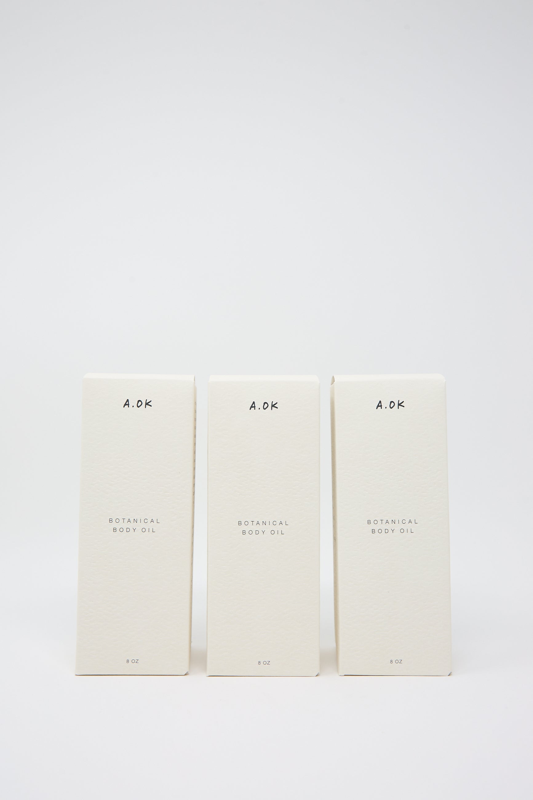 Three identical off-white boxes of Botanical Body Oil are arranged in a row against a plain white backdrop. The packaging features minimalist design and the text "A.OK Botanical Body Oil." This luxurious blend harnesses the power of natural oils, offering a refined unisex aroma.