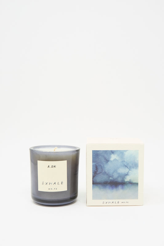 An "Exhale Candle" by A.OK, scented with soothing cardamom, is positioned beside a box adorned with a blue watercolor design on a simple white backdrop.