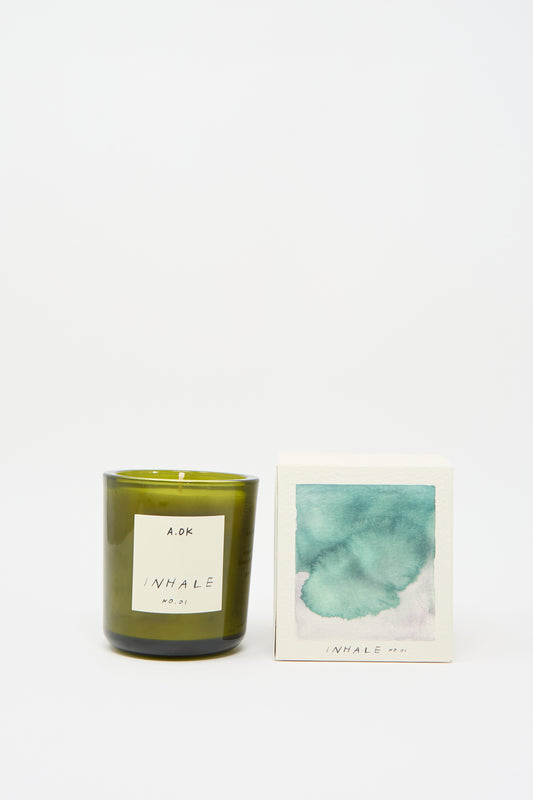 A green glass candle from A.OK, named "Inhale Candle," is placed next to a box decorated with a watercolor design and the same label, and it is infused with calming notes of vetiver.