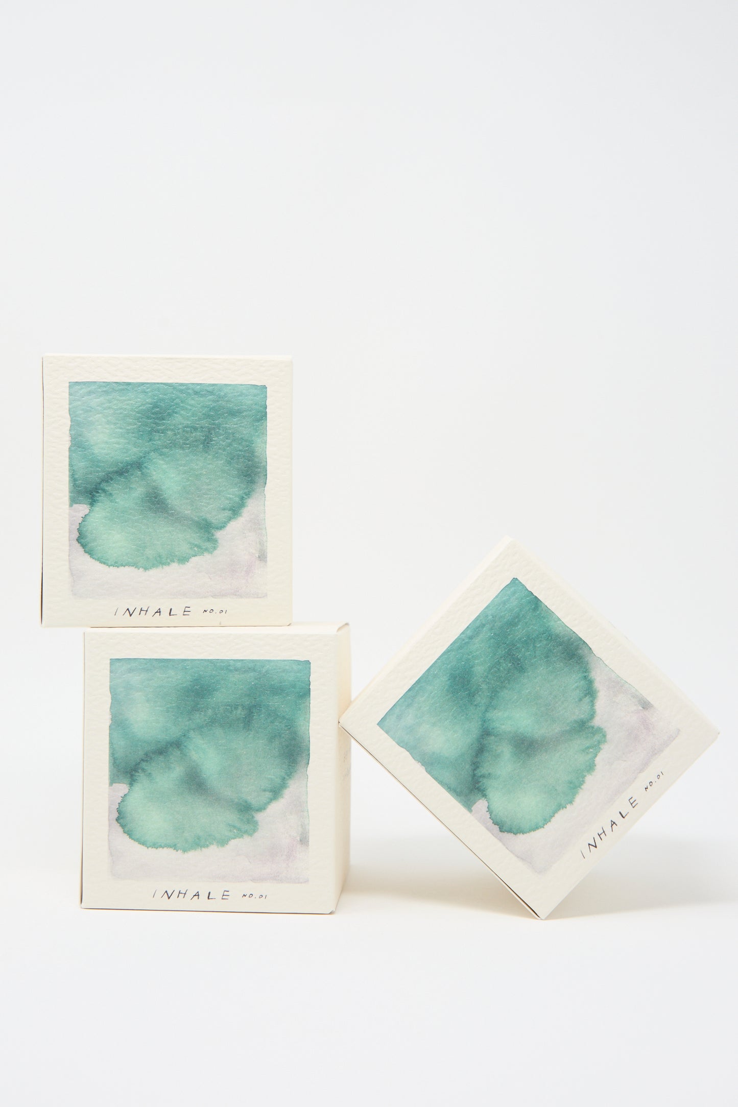 Three pastel art pieces in square frames, each labeled "Inhale Candle" from the A.OK brand, are stacked in a balanced arrangement against a plain white background, evoking the soothing scents of cedarwood and vetiver.