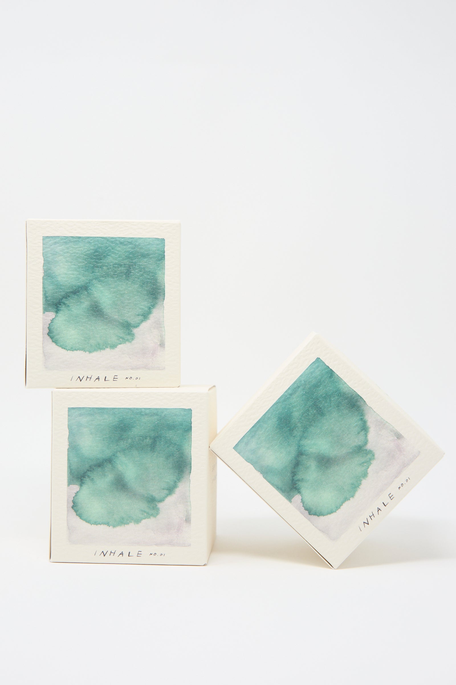 Three pastel art pieces in square frames, each labeled "Inhale Candle" from the A.OK brand, are stacked in a balanced arrangement against a plain white background, evoking the soothing scents of cedarwood and vetiver.