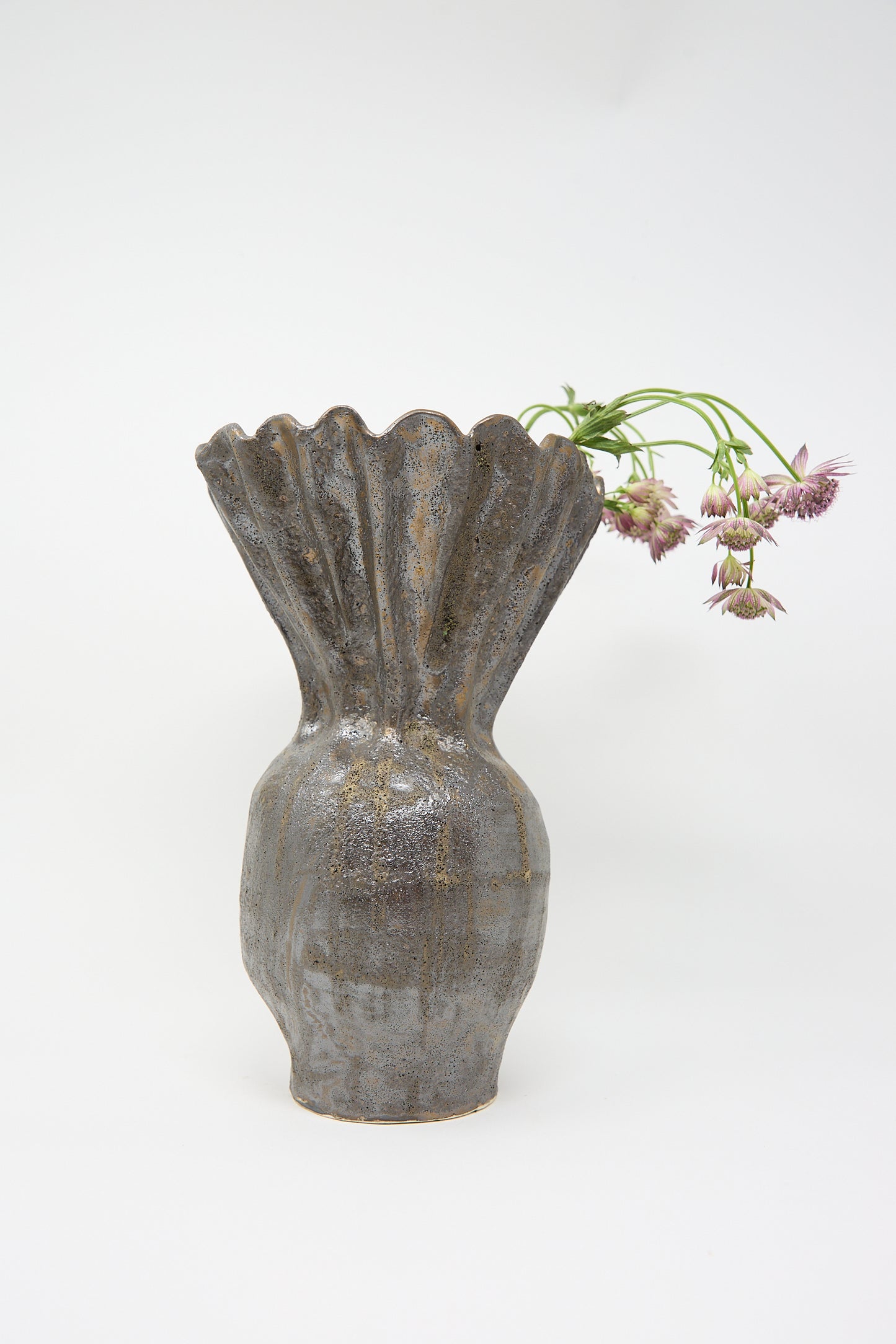 The Fold Vase in Bronze by ANK Ceramics, a tall stoneware piece with a textured finish and scalloped rim, holds wilting purple flowers against a plain background.