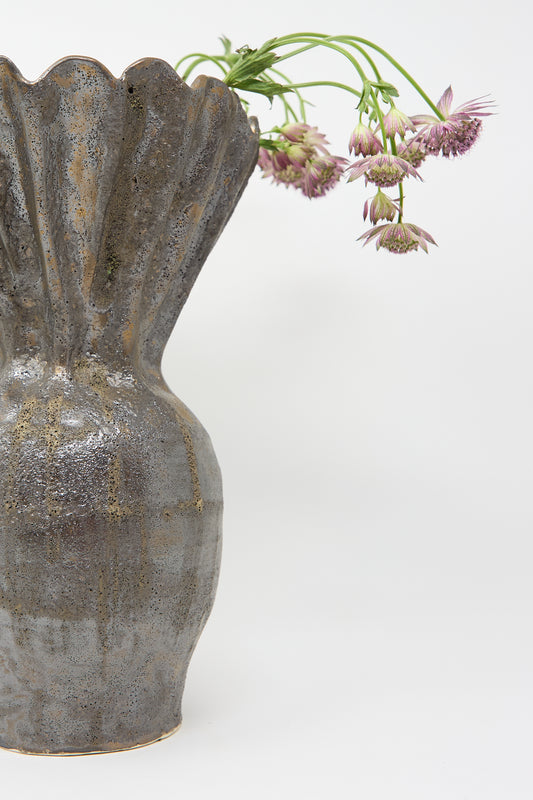 The Fold Vase in Bronze by ANK Ceramics, featuring a textured surface, beautifully displays drooping purple flowers against a plain white background.