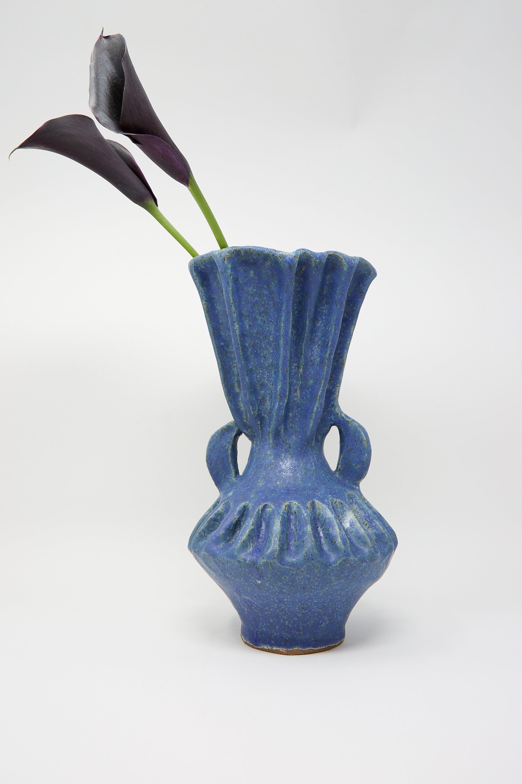 The Pleat Vase with Handles in Blue by ANK Ceramics showcases an exquisite fluted design complemented by a striking blue glaze and dual handles. This elegant stoneware vase beautifully contrasts two dark purple calla lilies against its light gray background.