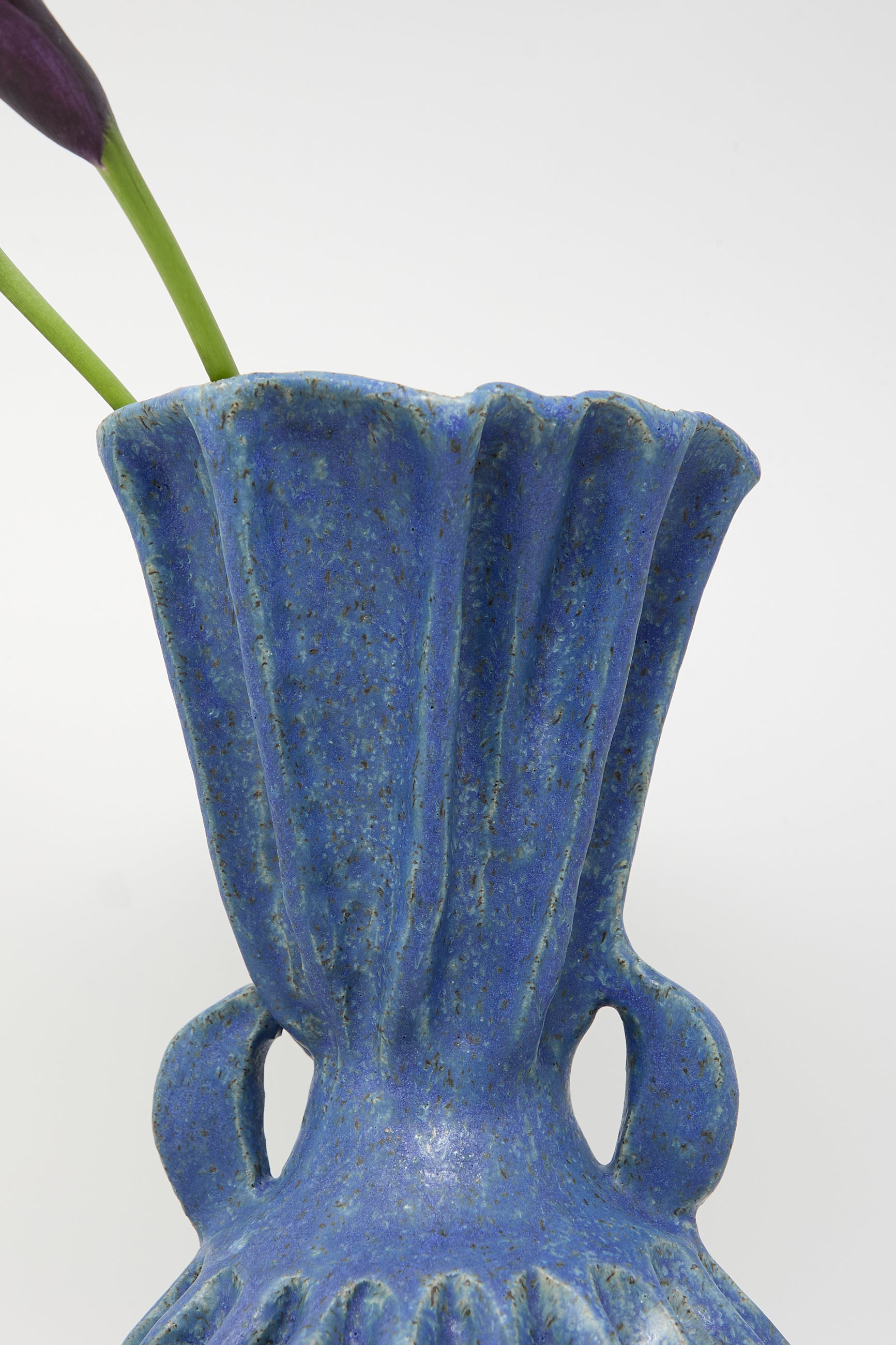 Close-up of an ANK Ceramics Pleat Vase with Handles in Blue, featuring a textured stoneware design with fluted details, containing a few green stems.