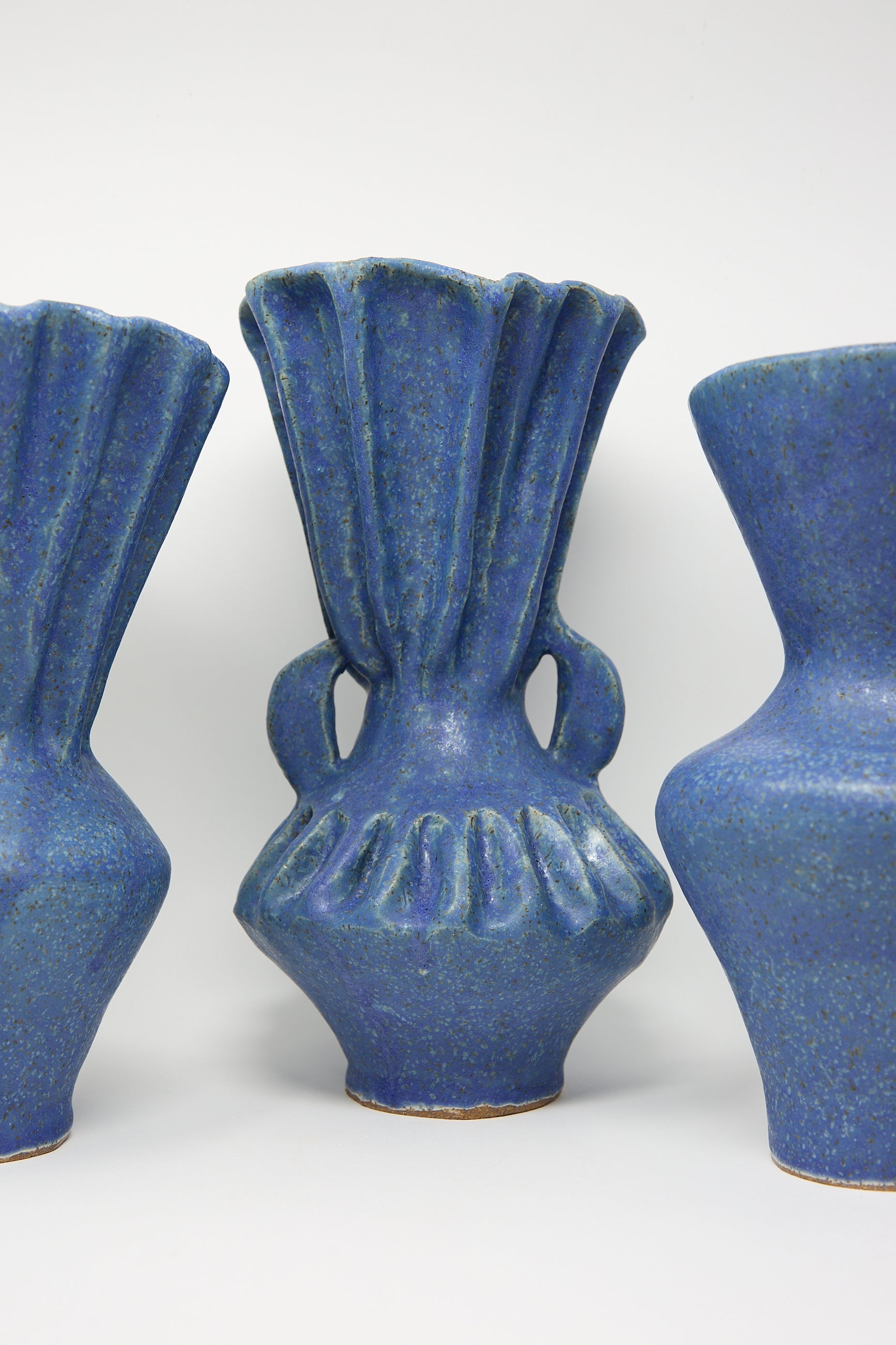 Three vases from ANK Ceramics stand elegantly on a plain background, all crafted from stoneware with a beautiful blue glaze. The center piece is the Pleat Vase with Handles in Blue, featuring a fluted design that sets it apart, while the surrounding vases display sleek, curved forms.