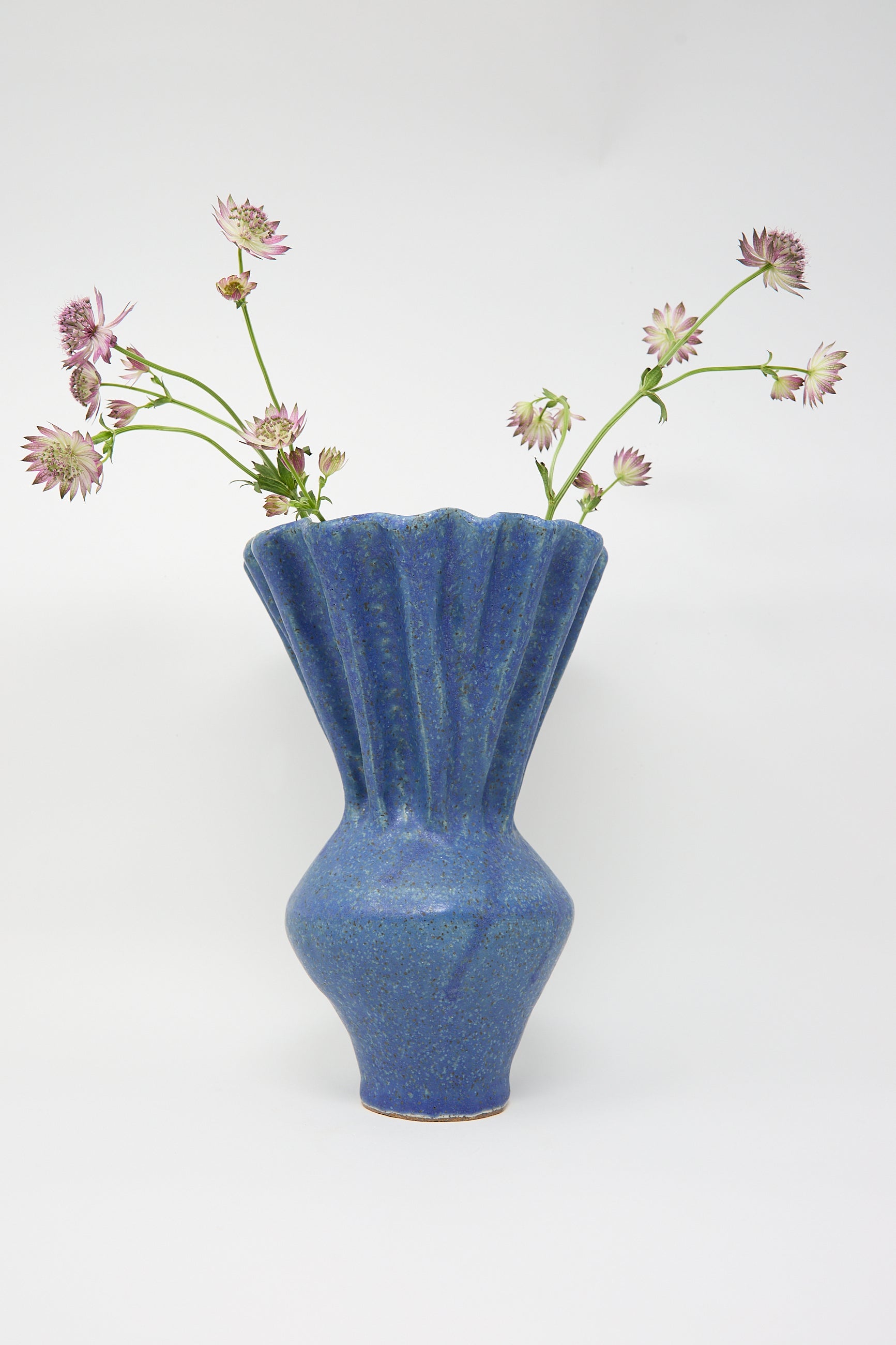 A Pleat Vase in Blue by ANK Ceramics, featuring a fluted design, is filled with several stems of small pink and white flowers against a plain white background.