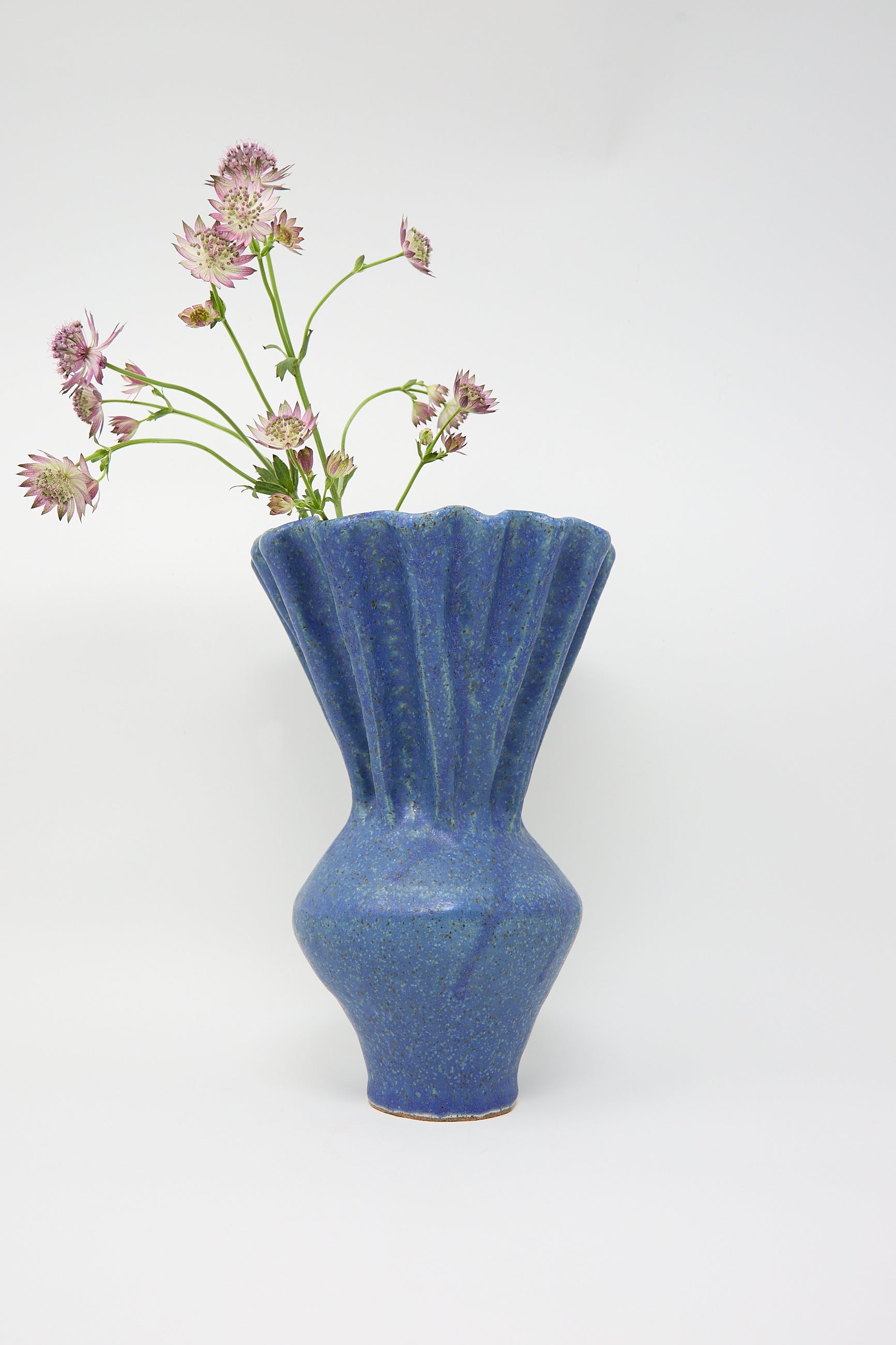 The Pleat Vase in Blue by ANK Ceramics elegantly holds a few pink flowers against a plain background.