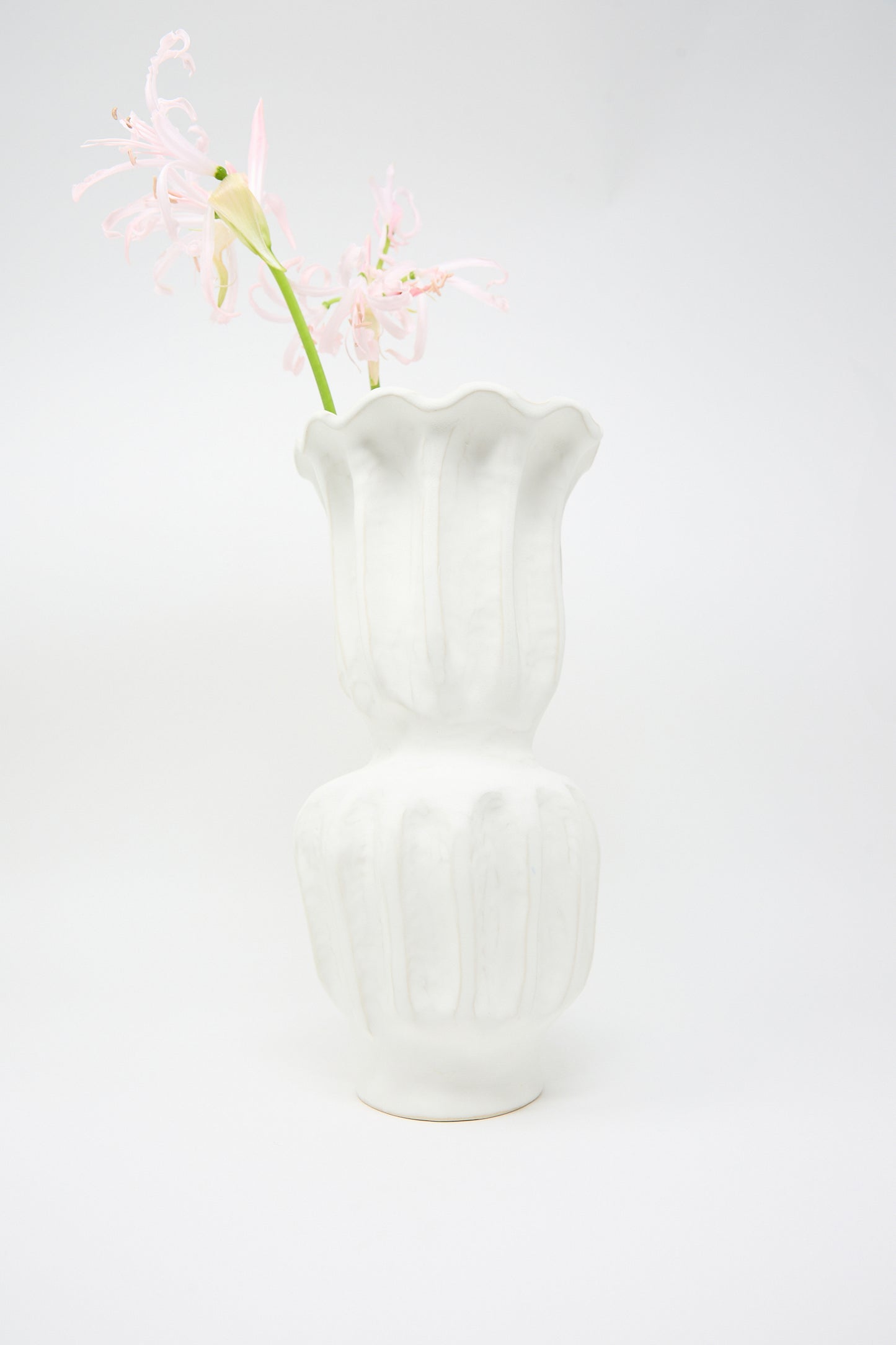 A single stem of light pink flowers stands out against a plain background in the Pleat Vase from ANK Ceramics, featuring a matte white glaze and an eye-catching wavy design.