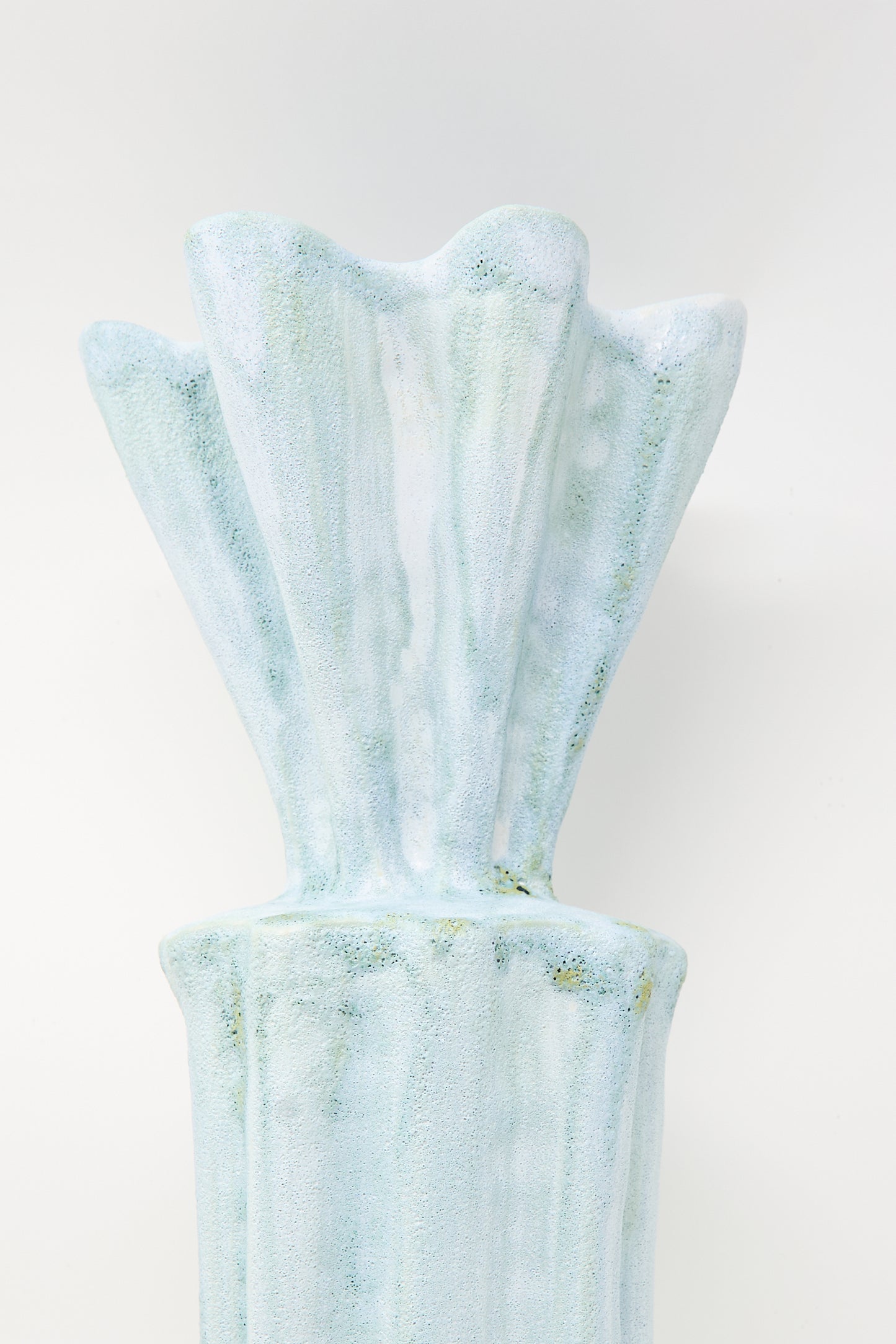 The "Tall Watercolor Vessel" by ANK Ceramics is a ceramic sculpture reminiscent of a fluted column, featuring a textured, pale blue and white surface set against a plain background.