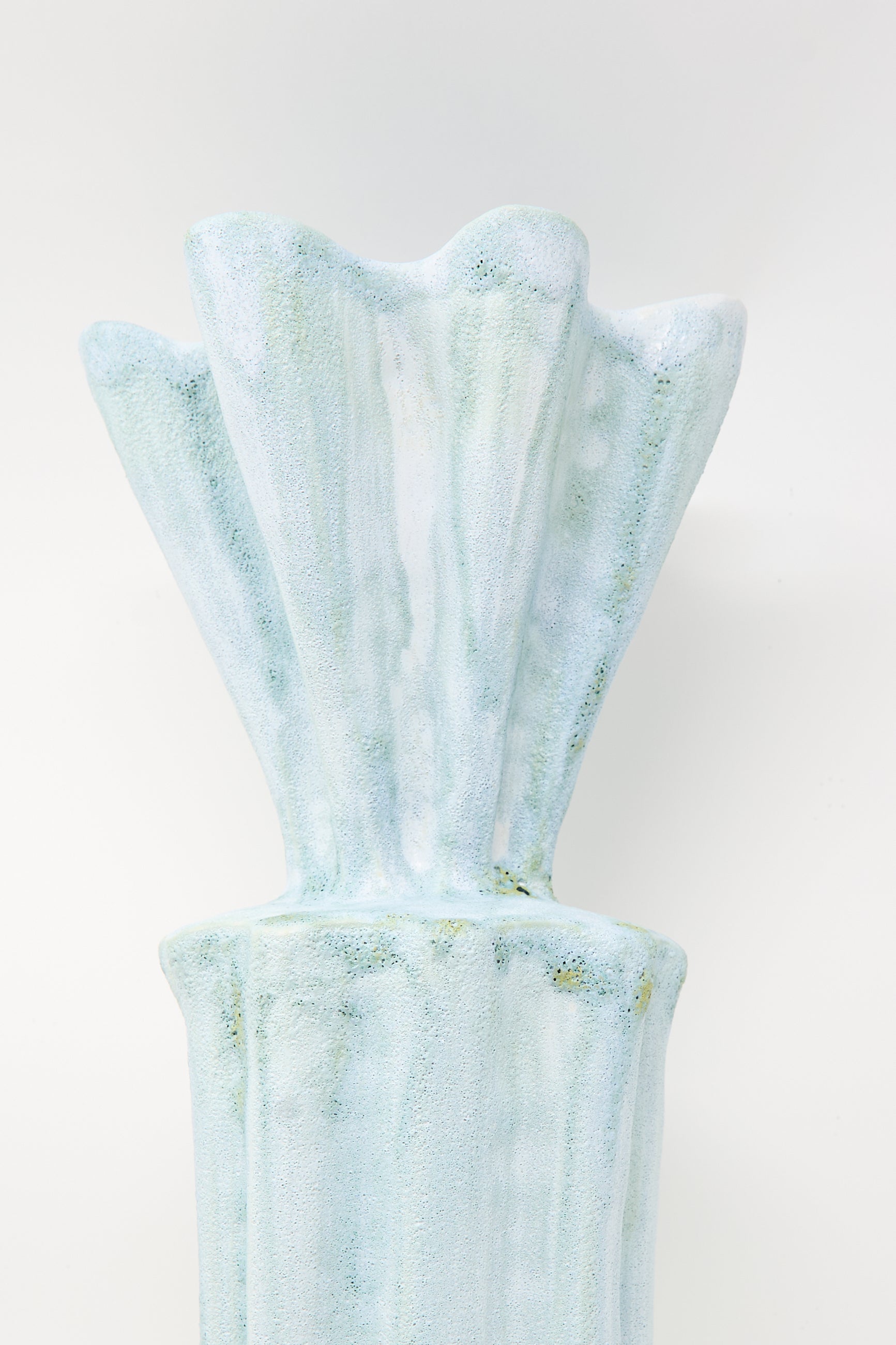 The "Tall Watercolor Vessel" by ANK Ceramics is a ceramic sculpture reminiscent of a fluted column, featuring a textured, pale blue and white surface set against a plain background.