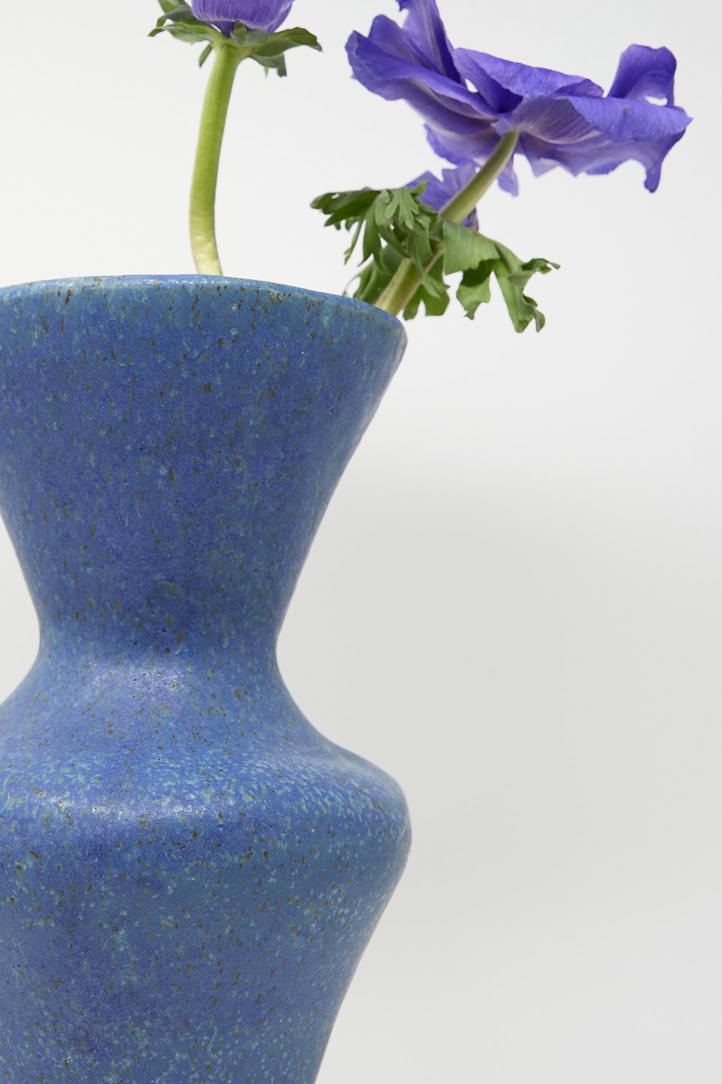 A close-up of the "Vase in Blue" by ANK Ceramics, featuring a blue glaze and adorned with a purple flower, set against a plain white background to beautifully showcase the charm of handcrafted ceramics.