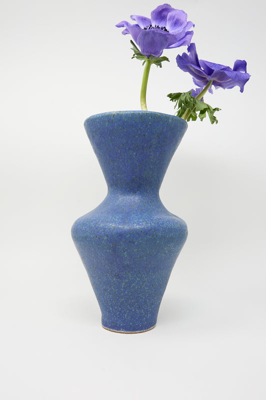 The Vase in Blue from ANK Ceramics, adorned with a blue glaze, holds two purple flowers with green stems against a simple white backdrop.