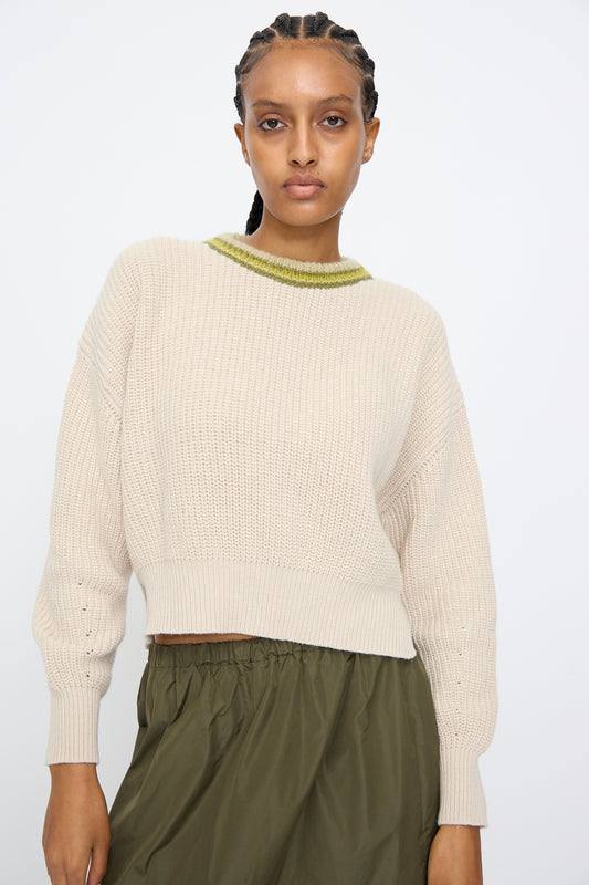 A woman wears the AVN American Sweater in Sand with a green collar detail and an olive green skirt, standing against a plain white background.
