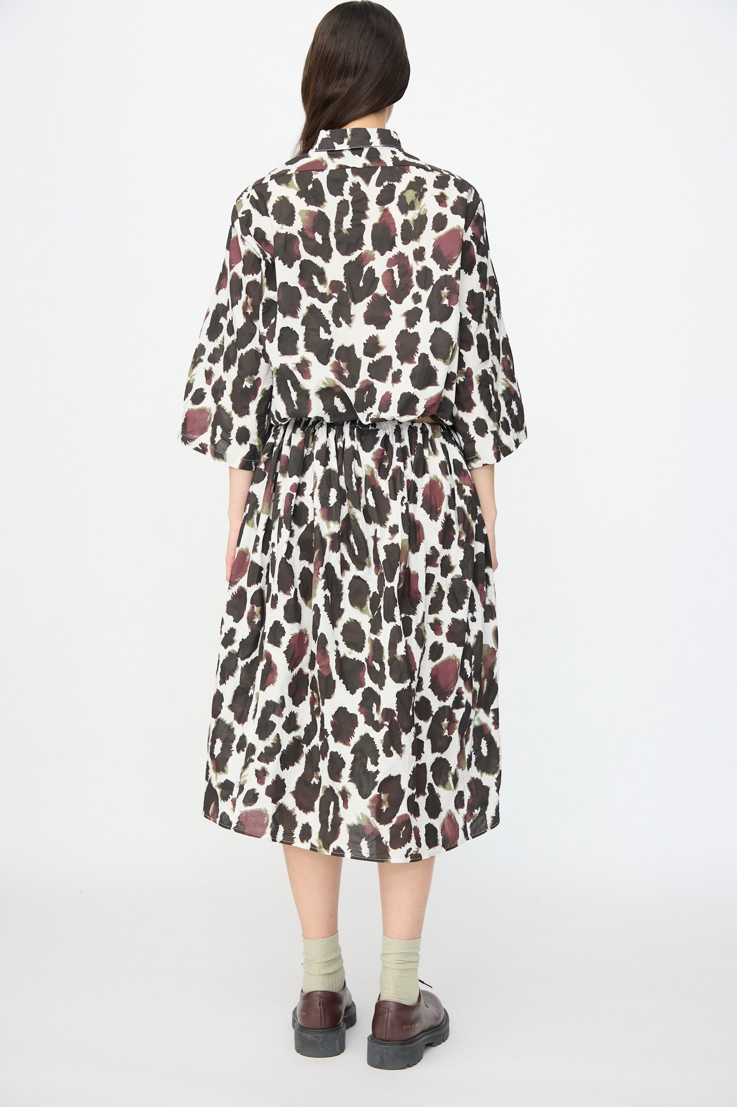 A person stands with their back to the camera, wearing the AVN Bloom Skirt in White with Print styled as a dress, paired with light socks and brown shoes.