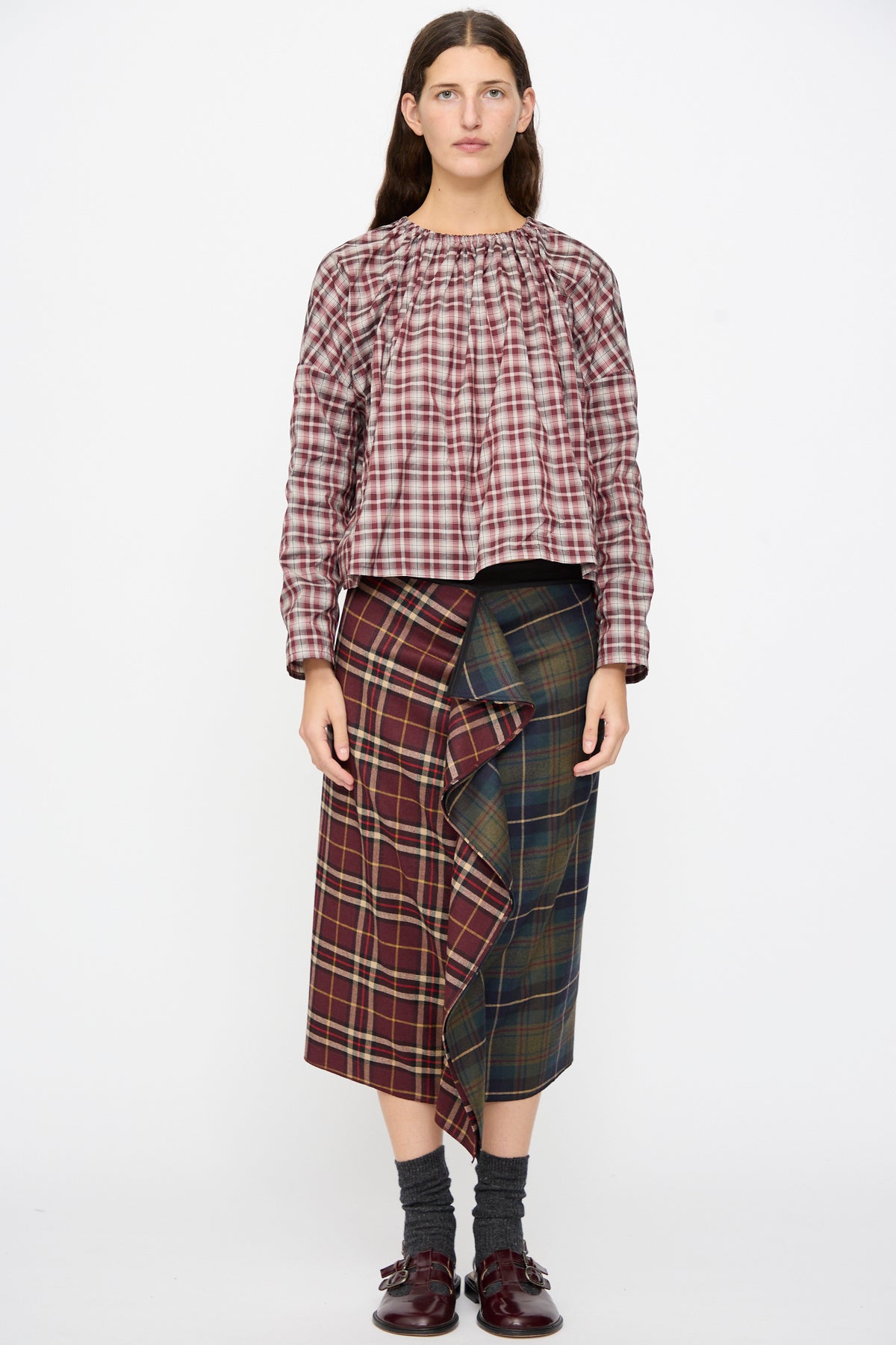 A person wearing a bordeaux check Bolla Shirt with a ruffled neck and boxy fit, paired with a multi-patterned plaid midi skirt with a ruffled front, dark socks, and buckled shoes.