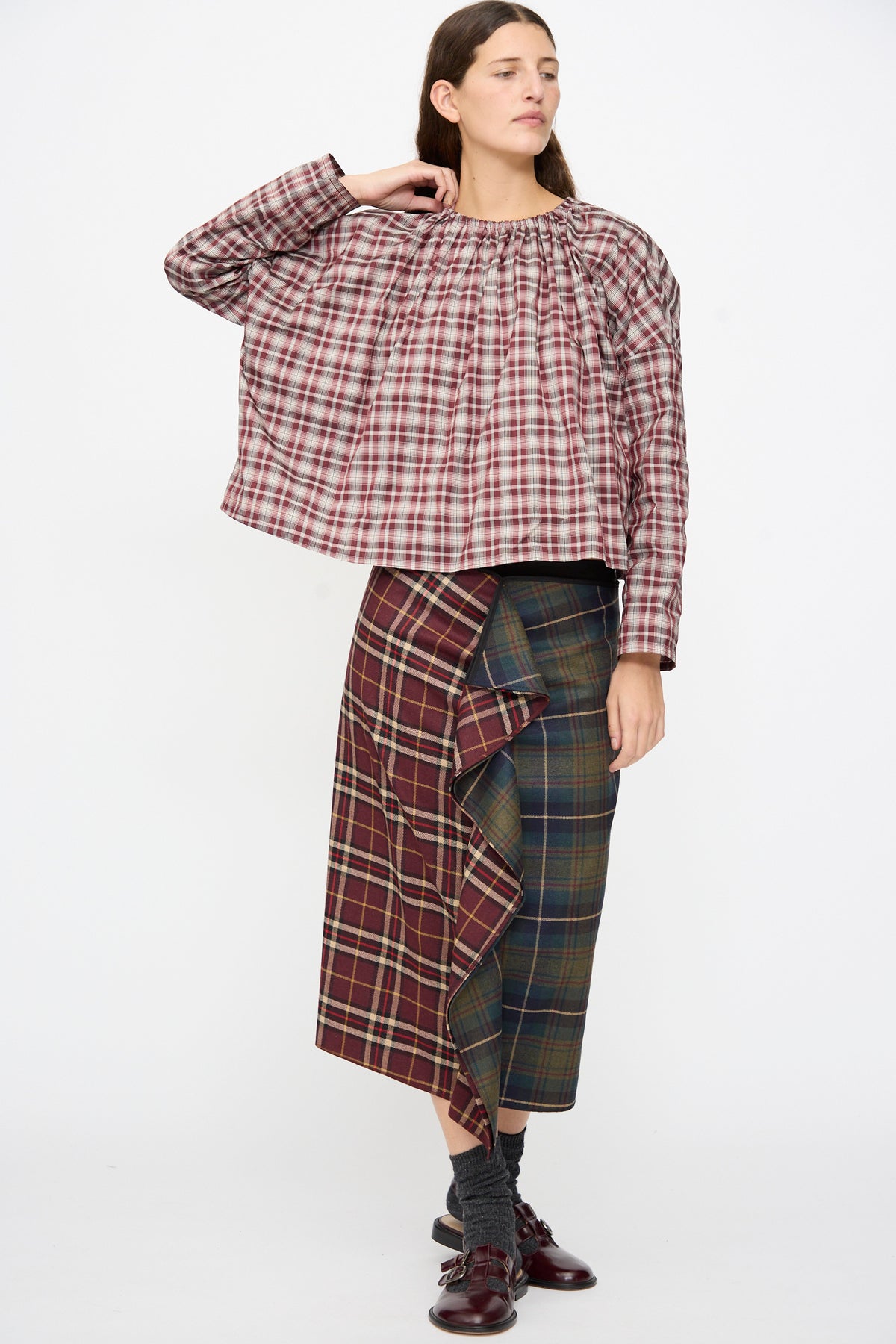 A person wearing a boxy fit, long-sleeved Bolla Shirt in Bordeaux Check and a knee-length, multi-patterned plaid skirt with a ruffled detail, styled with dark socks and buckled shoes.