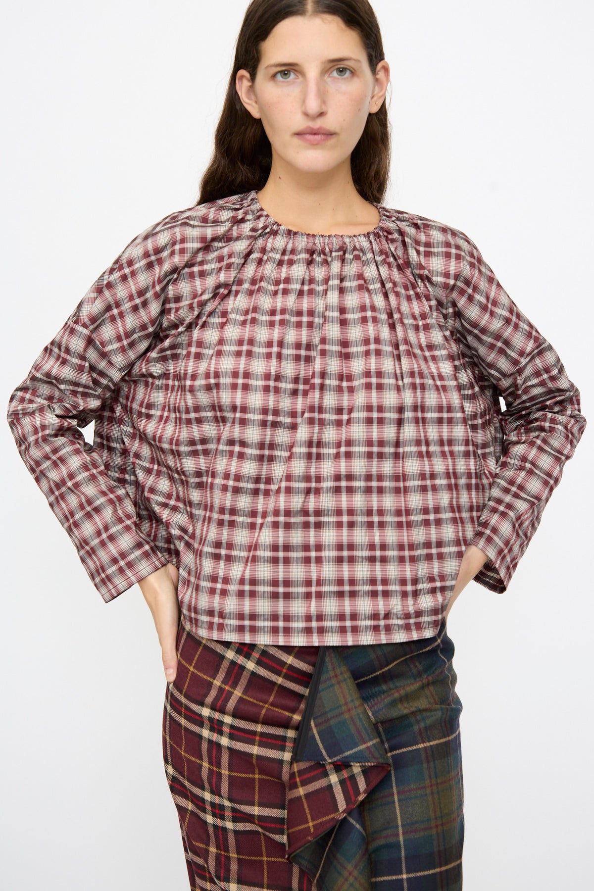 A person wearing a boxy-fit, maroon-and-white Bolla Shirt in Bordeaux Check with a gathered neckline, paired with a pleated, multi-patterned plaid skirt.