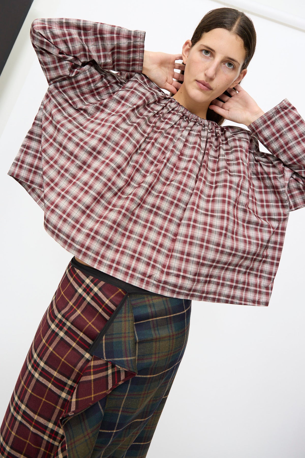 A person wearing an AVN Bolla Shirt in Bordeaux Check, paired with a multi-patterned plaid skirt with a ruffled front.