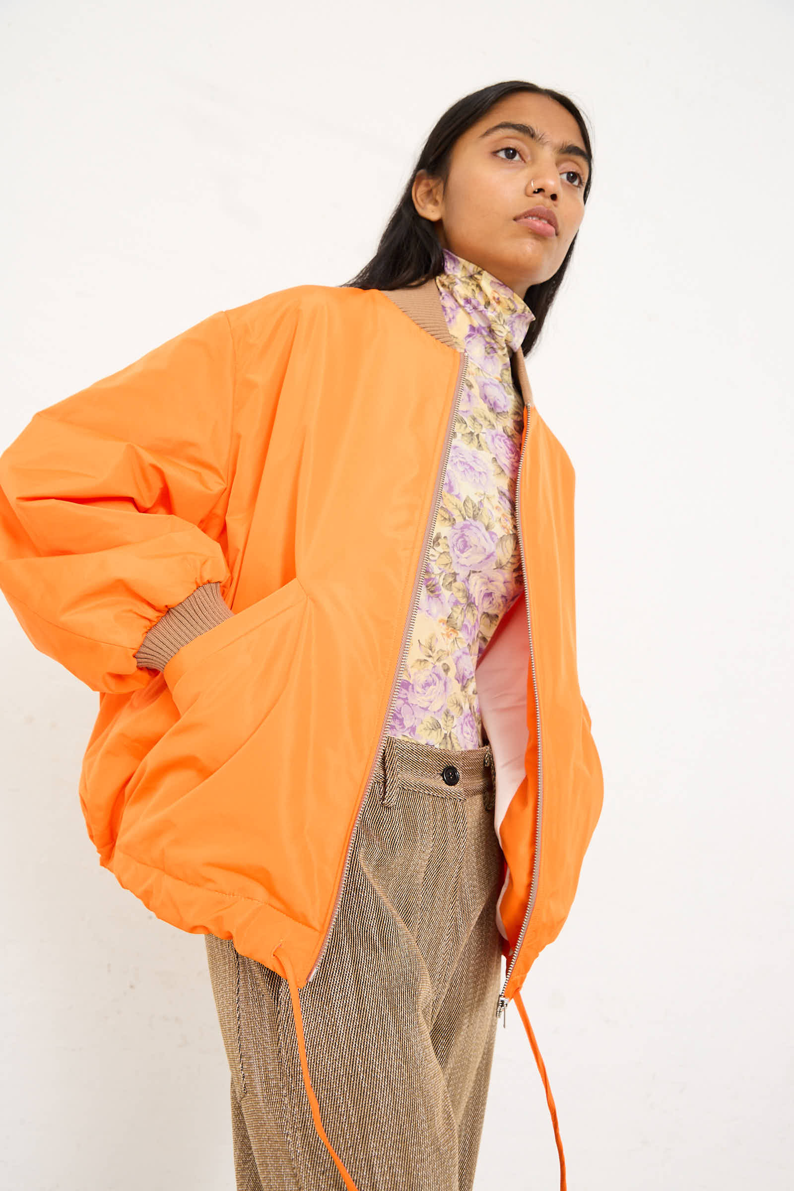 Person wearing an oversized fit AVN Bomber Jacket in Orange over a floral top and beige pants, standing against a plain background.