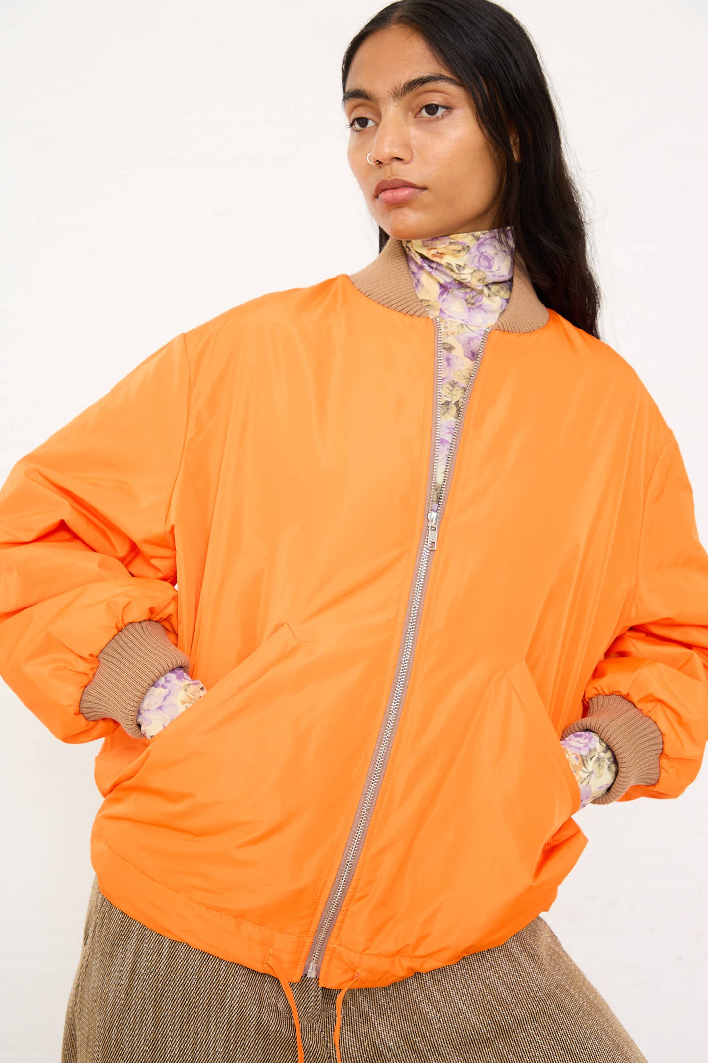 Person wearing an AVN Bomber Jacket in Orange, designed with an oversized fit taffeta material, layered over a floral top, standing with hands in pockets.