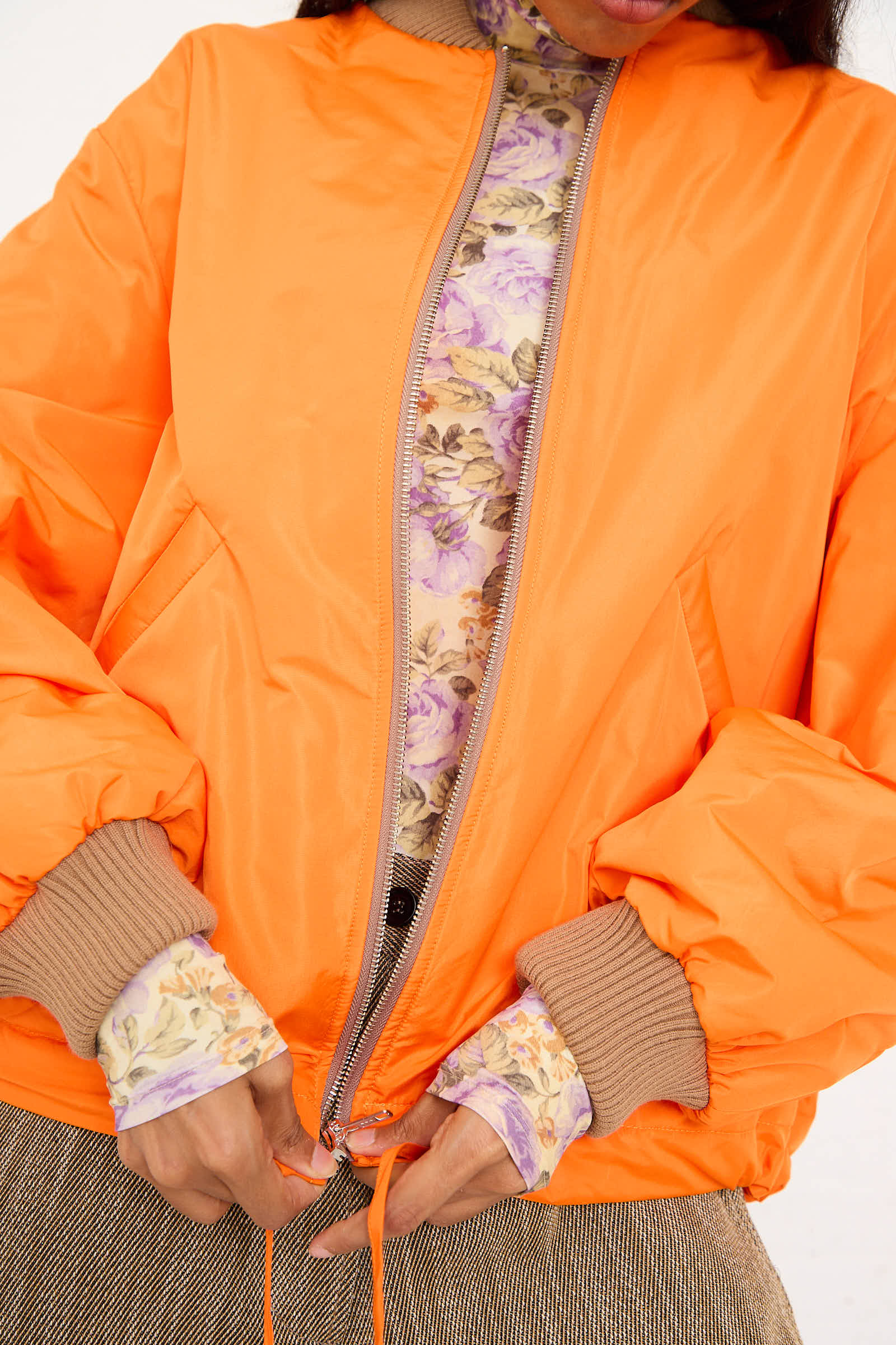 A person zips up an AVN Bomber Jacket in Orange. They are wearing a floral shirt underneath and brown pants.