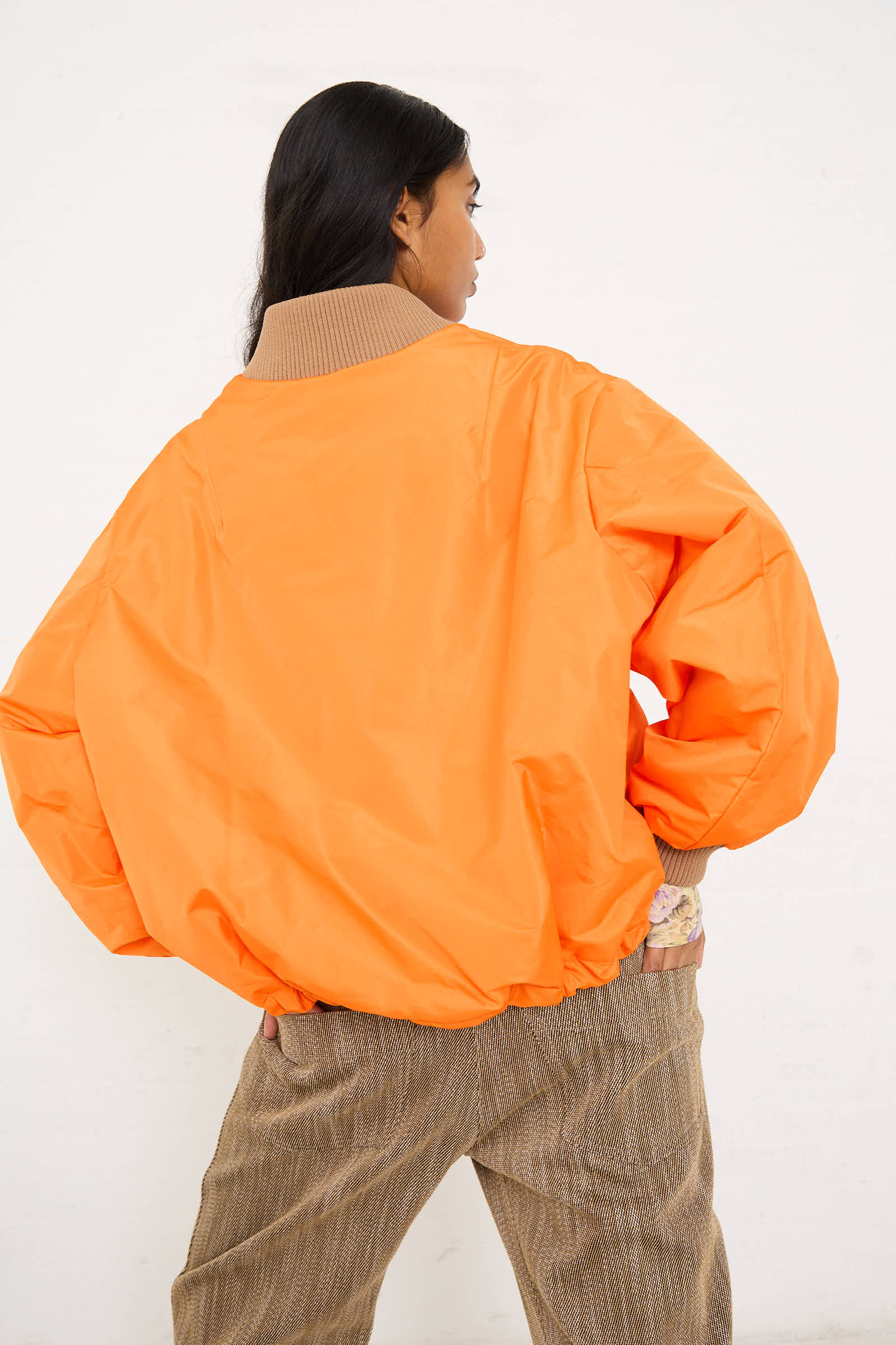 Person with long dark hair wearing an oversized fit Bomber Jacket in Orange by AVN and brown pants, standing with back to the camera.