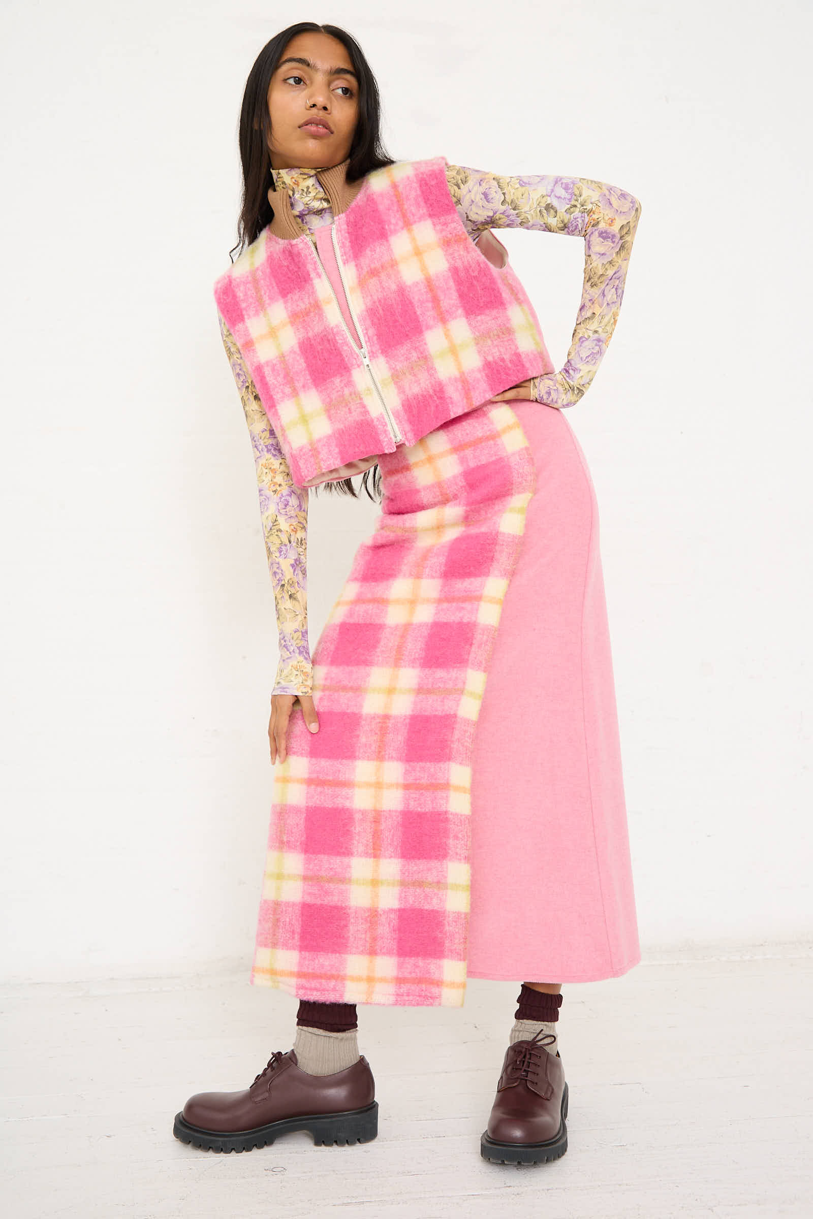 A person stands against a white wall, wearing an AVN Crossed Skirt in White, Pink and Green Check over a matching plaid maxi skirt and plain pink long skirt, with a floral long-sleeve shirt underneath and brown shoes.