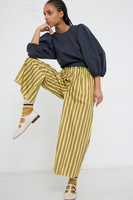 A woman in a dark blouse and AVN Italian womenswear Easy Pant in Brown and Yellow Stripes posing with one hand on her face and one knee bent, standing against a white background.