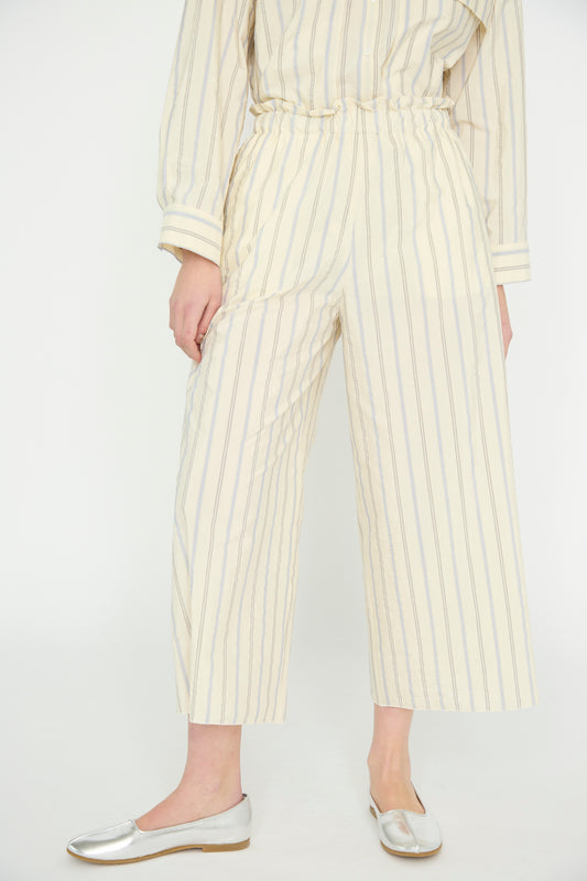 A model is dressed in the AVN Easy Pant in Off White with Stripe and a matching off-white, cotton blend long-sleeve shirt, complemented by silver ballet flats.